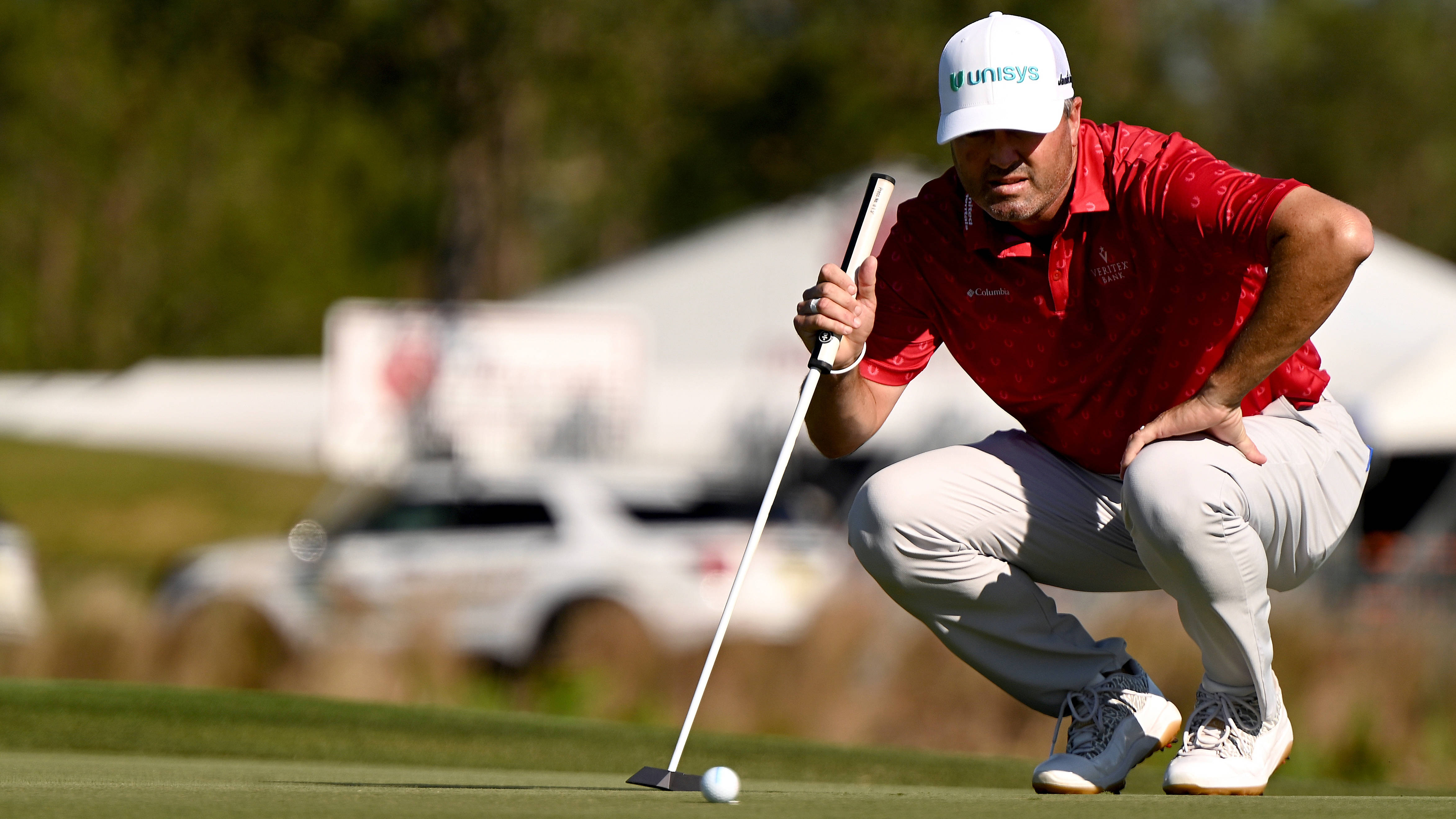 Ryan Palmer 桌布 壁紙 — 一位身穿紅色襯衫的高爾夫球手在陽光明媚的果嶺上專注地蹲著推桿，背景是模糊的觀眾和汽車。
