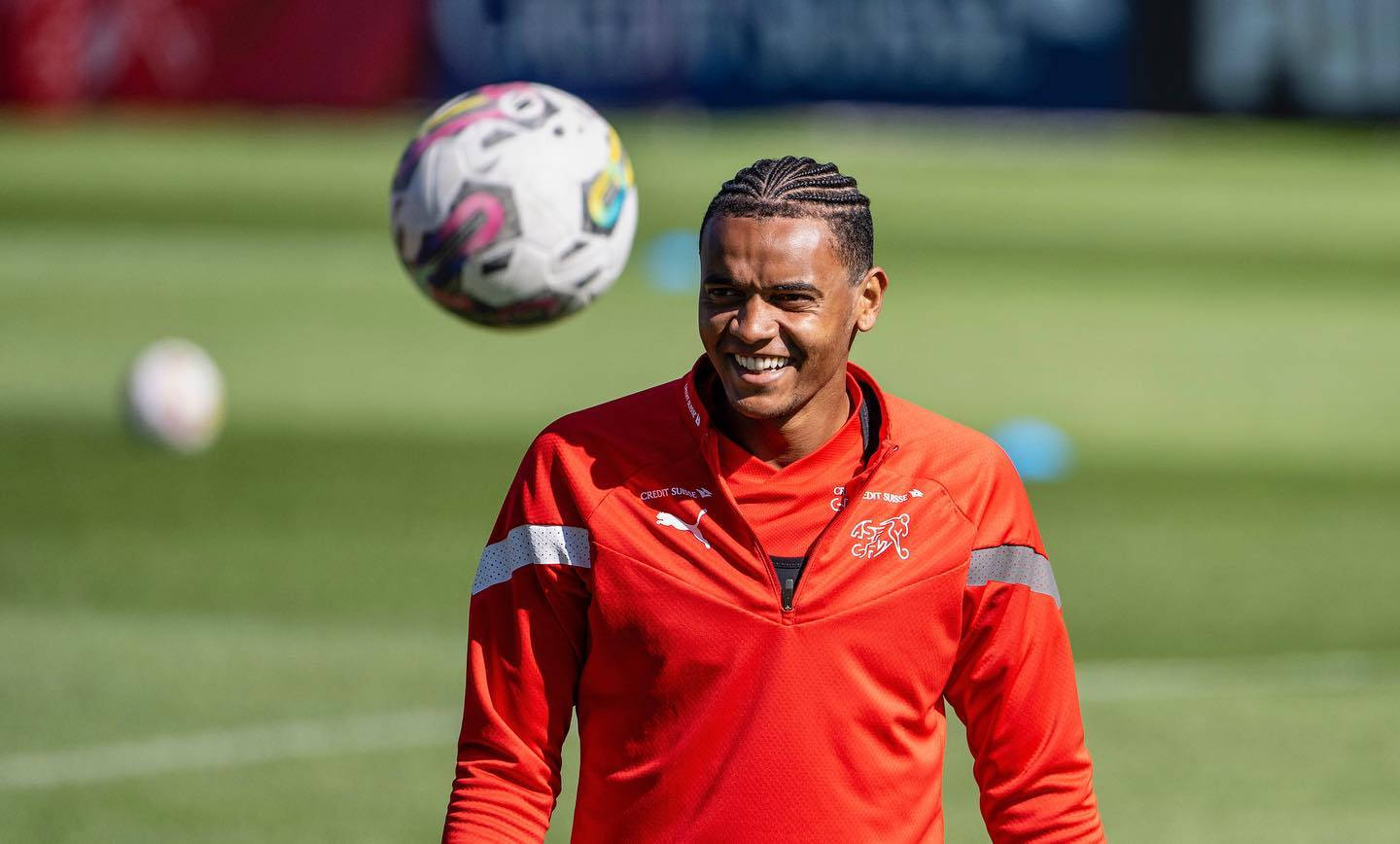 Soccer Ball 桌布 壁紙 — 一位身穿紅色訓練外套、面帶微笑的足球運動員，在晴朗的白天於明亮的綠色球場上頂著一個色彩繽紛的球。