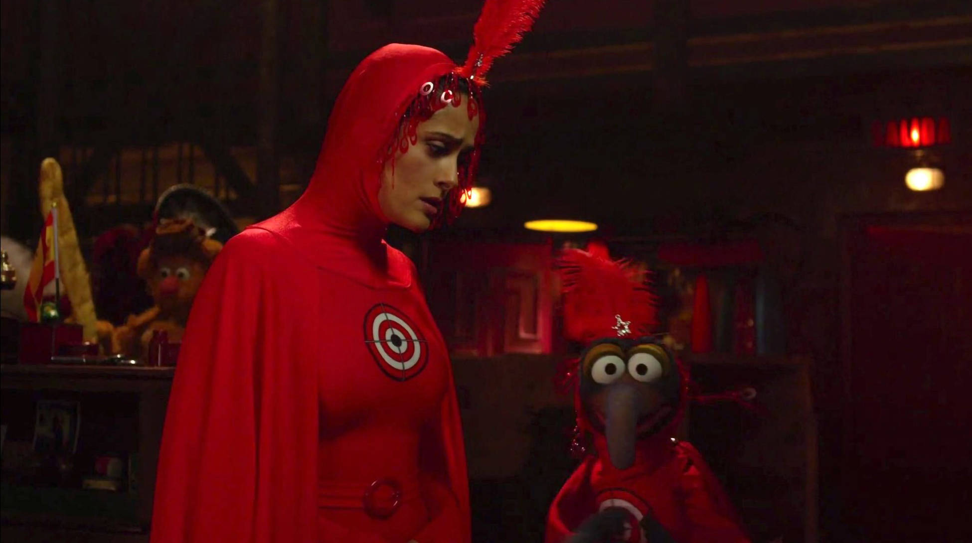 muppets 桌布 壁紙 — 一位身穿紅色連帽服飾、頭戴羽毛頭飾的女性，站在一個昏暗的房間裡，旁邊是一個紅色羽毛木偶。