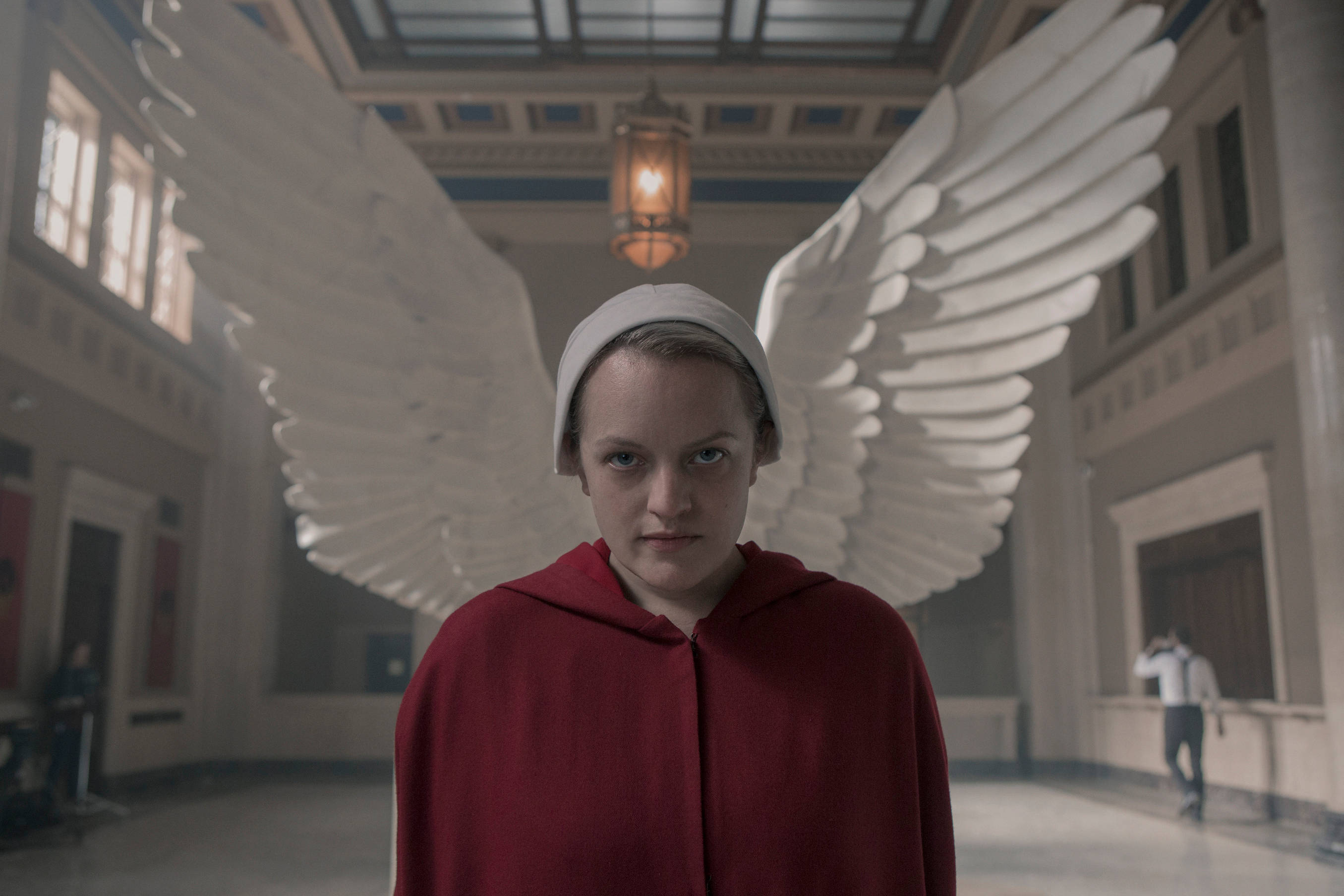 Hulu 桌布 壁紙 — 一位身穿紅色連帽長袍、擁有白色翅膀的女性，莊嚴地站在懸掛燈籠下宏偉的機構走廊中。
