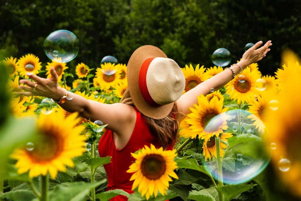 Beautiful Sunflower 桌布 壁紙 — 一位身穿紅色連衣裙和草帽的女性，在向日葵田中歡快地張開雙臂，泡泡在她周圍飄浮。