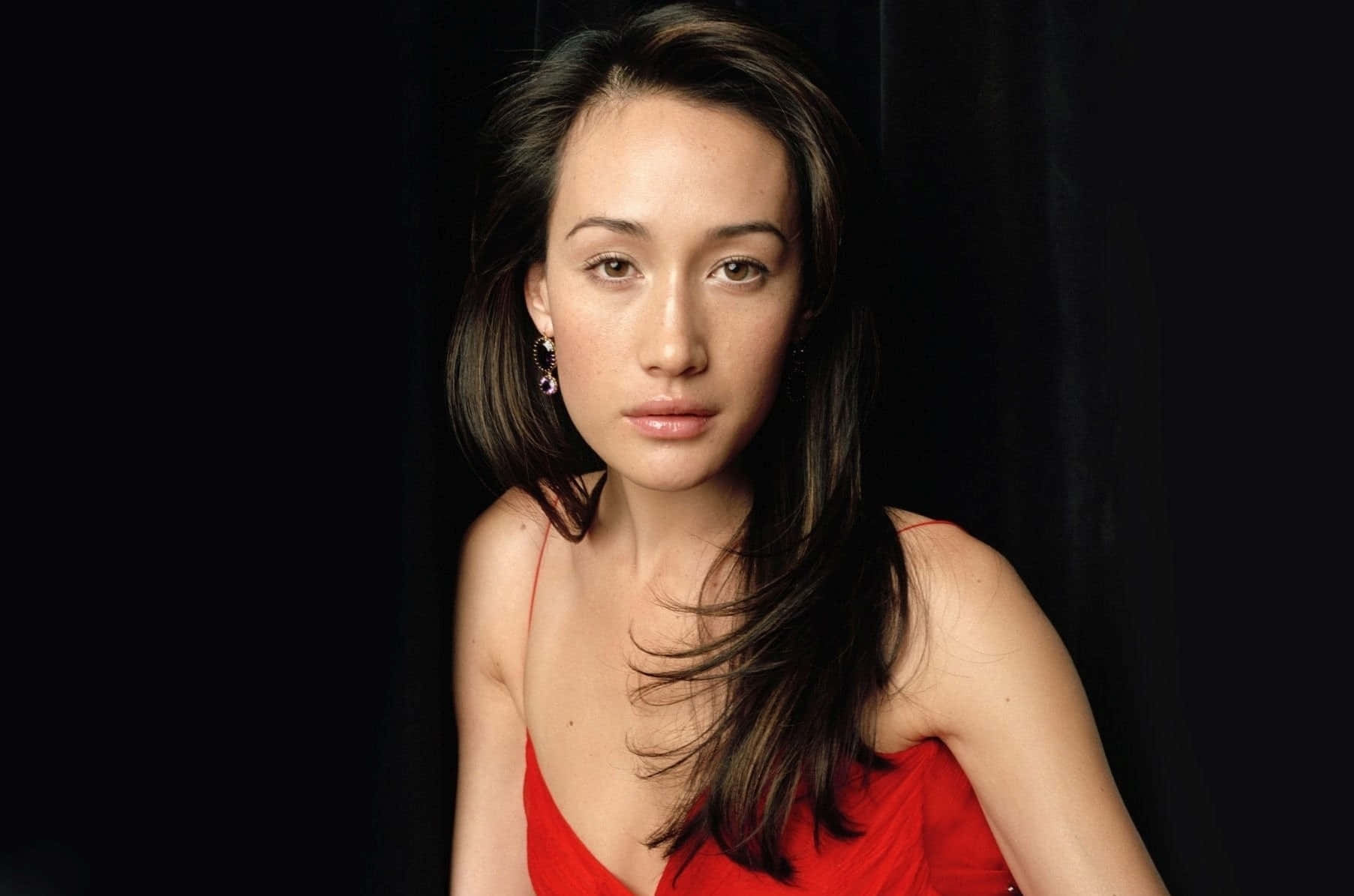 maggie q 桌布 壁紙 — 一位身穿紅色連衣裙的女性在 stark 黑色背景下柔和地凝視，微妙的燈光凸顯她的特徵。
