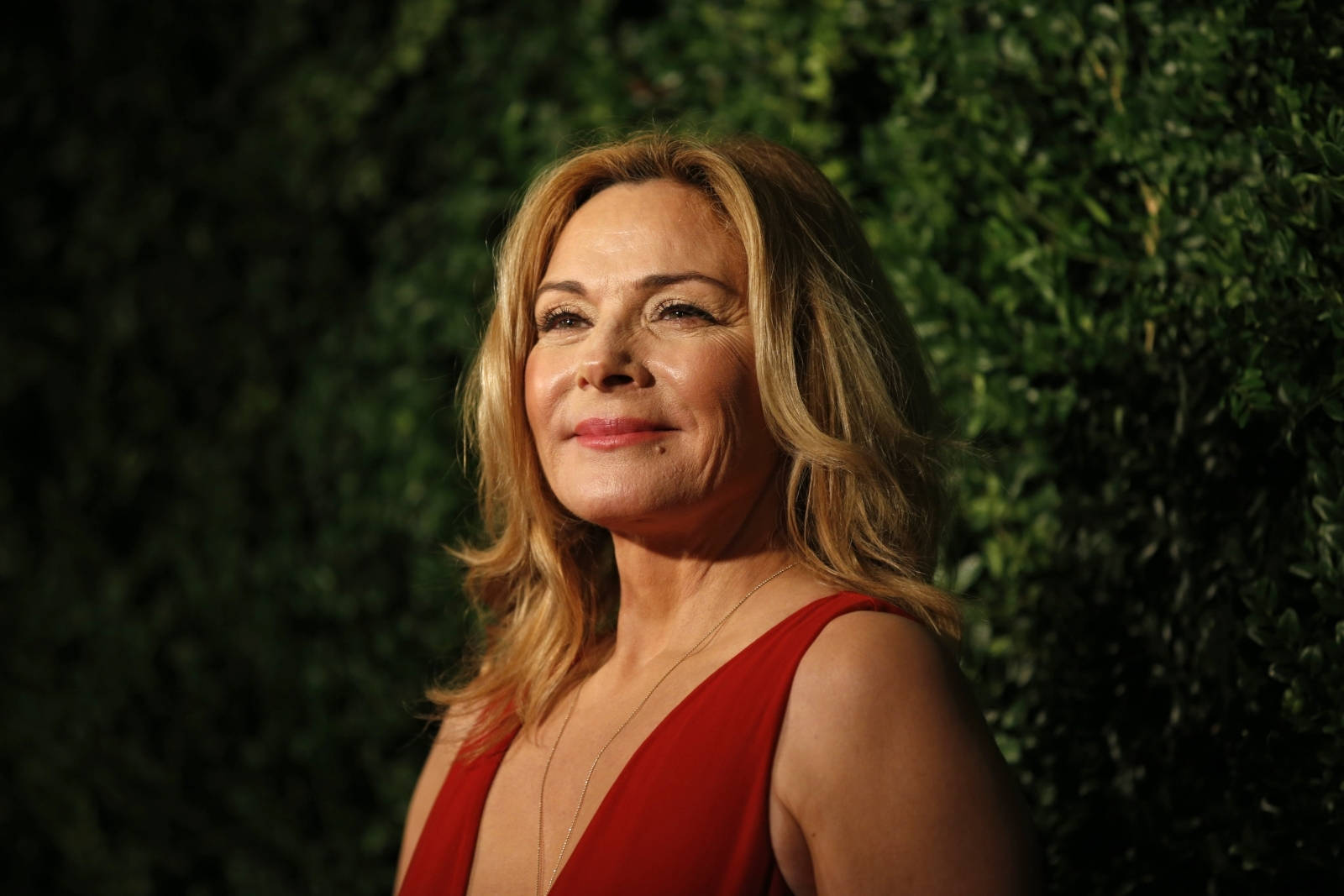 Celebrity 桌布 壁紙 — 一位身穿紅色連衣裙的女性在溫暖的燈光下，對著茂密的綠色樹籬背景溫柔地微笑。