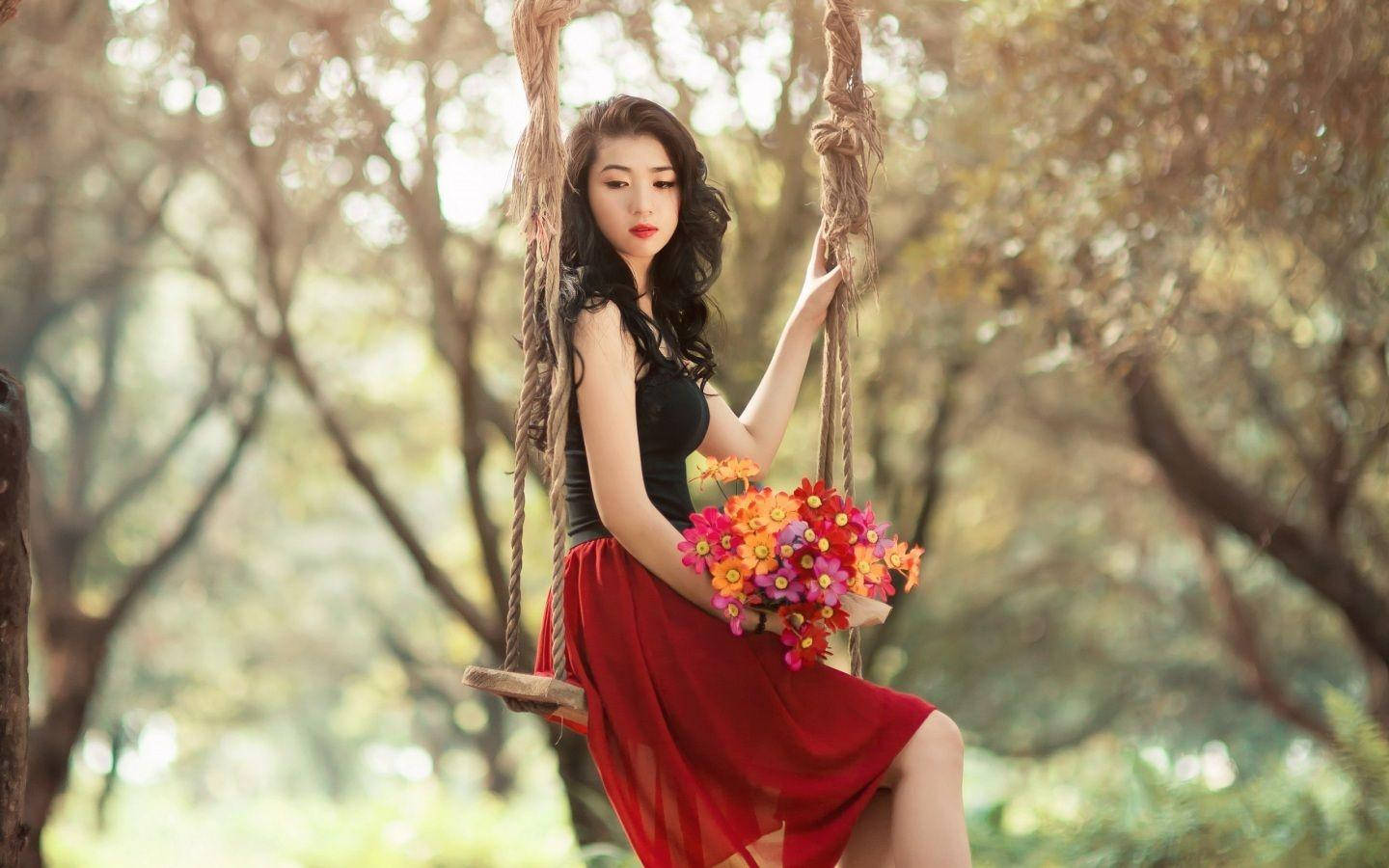 美女 桌布 壁紙 — 一位身穿紅色連衣裙的女性坐在繩索鞦韆上，在陽光明媚的森林中手持一束鮮豔的花束。