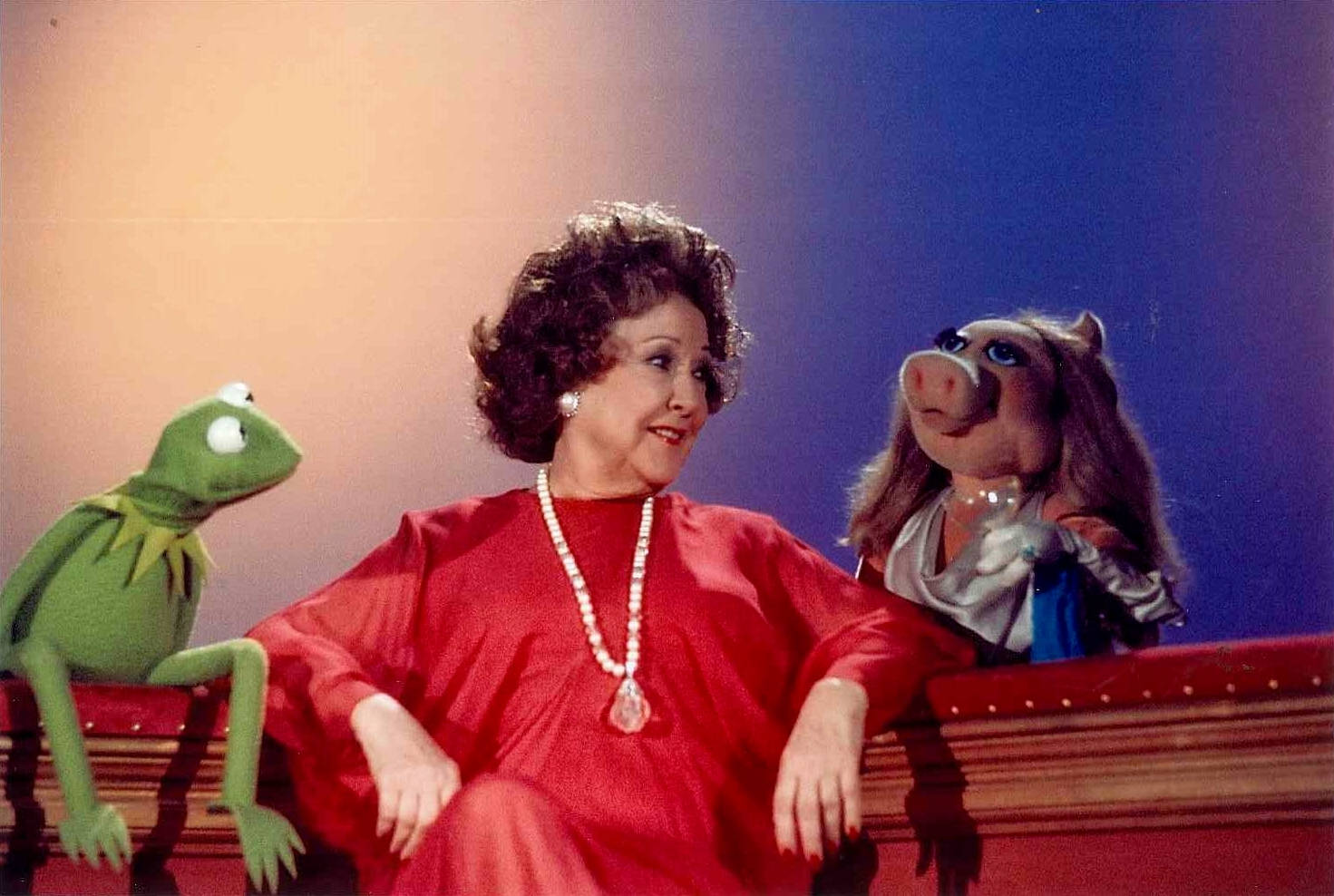 muppets 桌布 壁紙 — 一位身穿紅色連衣裙的女性坐在青蛙柯密特和豬小姐之間的舞台上，舞台上有溫暖的漸變燈光。