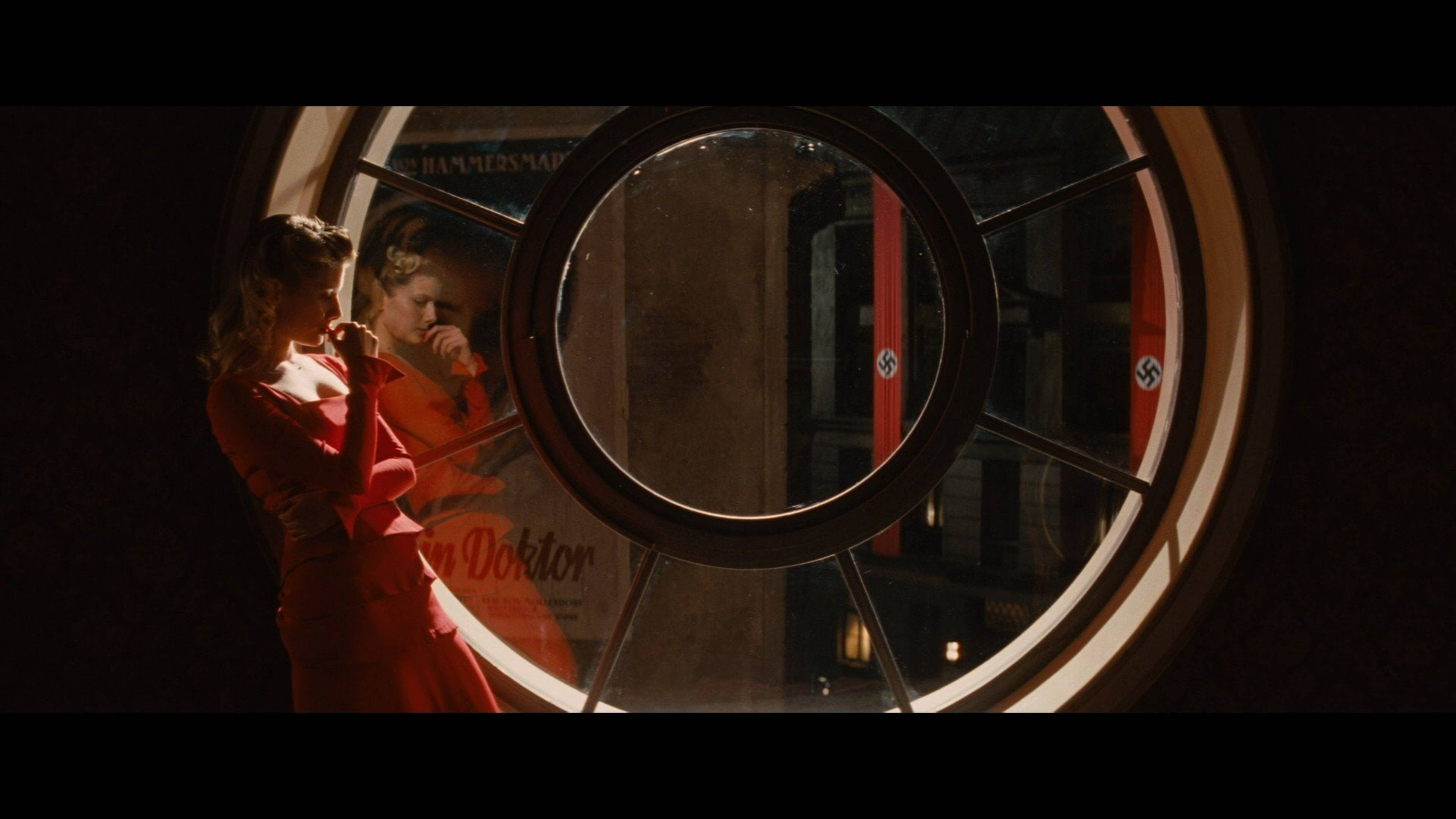Inglourious Basterds 桌布 壁紙 — 一位身穿紅色連衣裙的女性站在一個圓窗旁，窗戶反射出一條燈光昏暗、掛有卐字旗幟的街道。