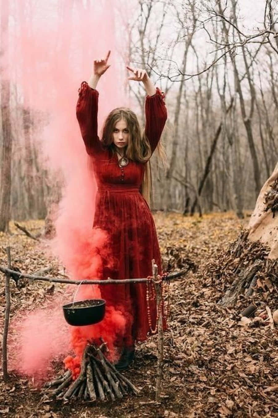 Witch Costumes 桌布 壁紙 — 一位身穿紅色連衣裙的女性站在光禿禿的森林中，粉紅色的煙霧從火上的大鍋中滾滾而出。