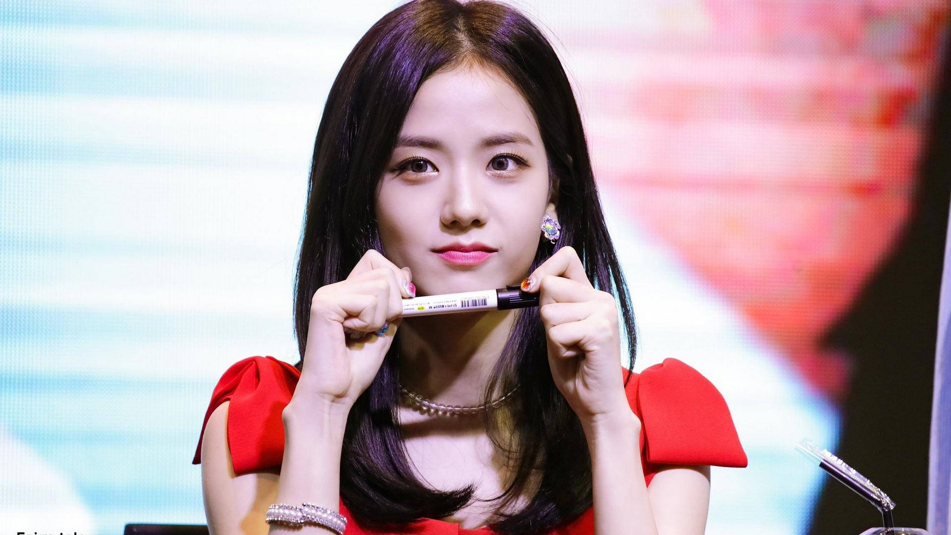 jisoo 可愛 桌布 壁紙 — 一位身穿紅色連衣裙的年輕女性，在模糊多彩的背景前，帶著俏皮的表情將記號筆靠近嘴唇。