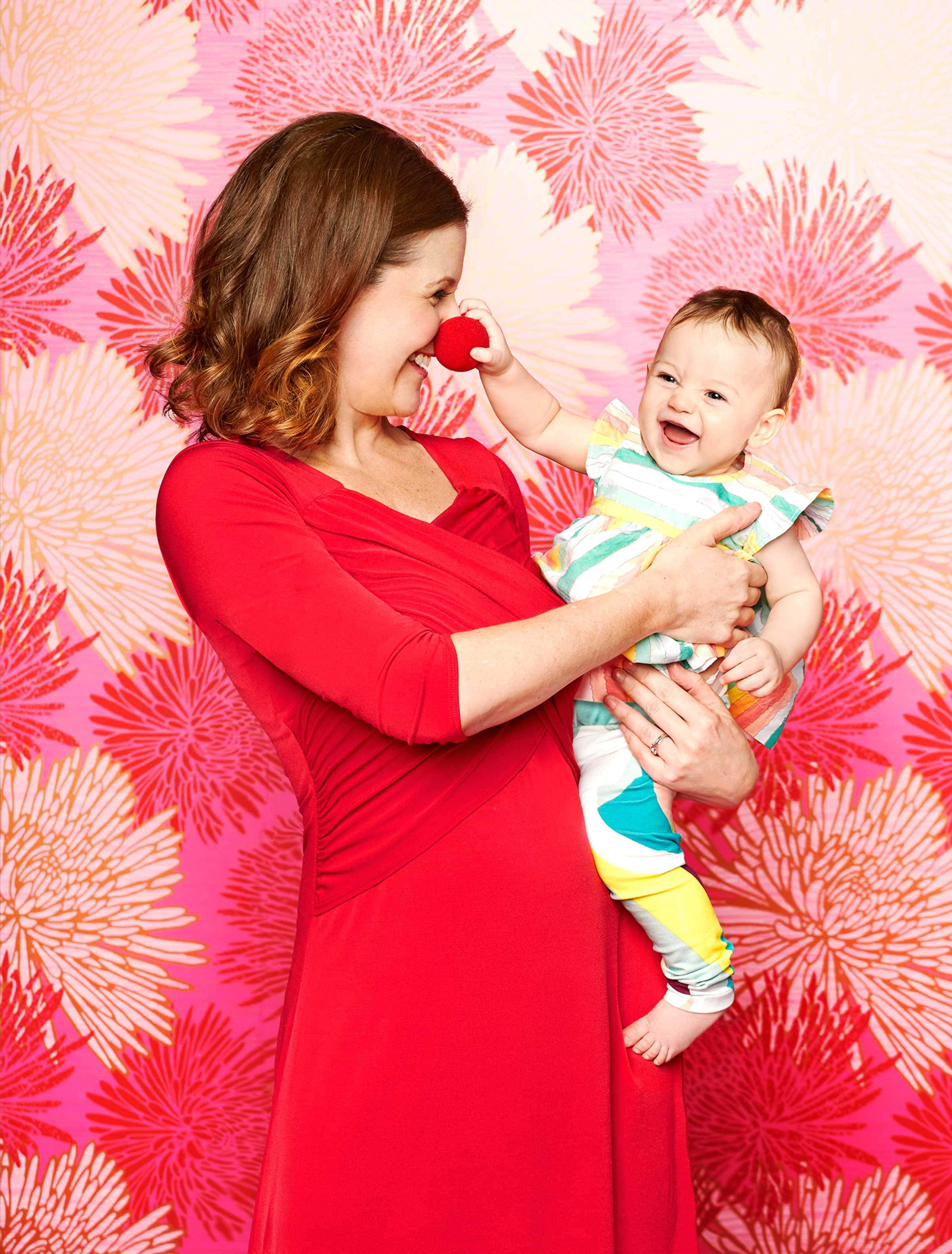 Family 桌布 壁紙 — 一位身穿紅色連衣裙的快樂母親大笑，她的寶寶頑皮地將一個紅色球碰到她的鼻子上，背景是充滿活力的粉紅色花卉壁紙。