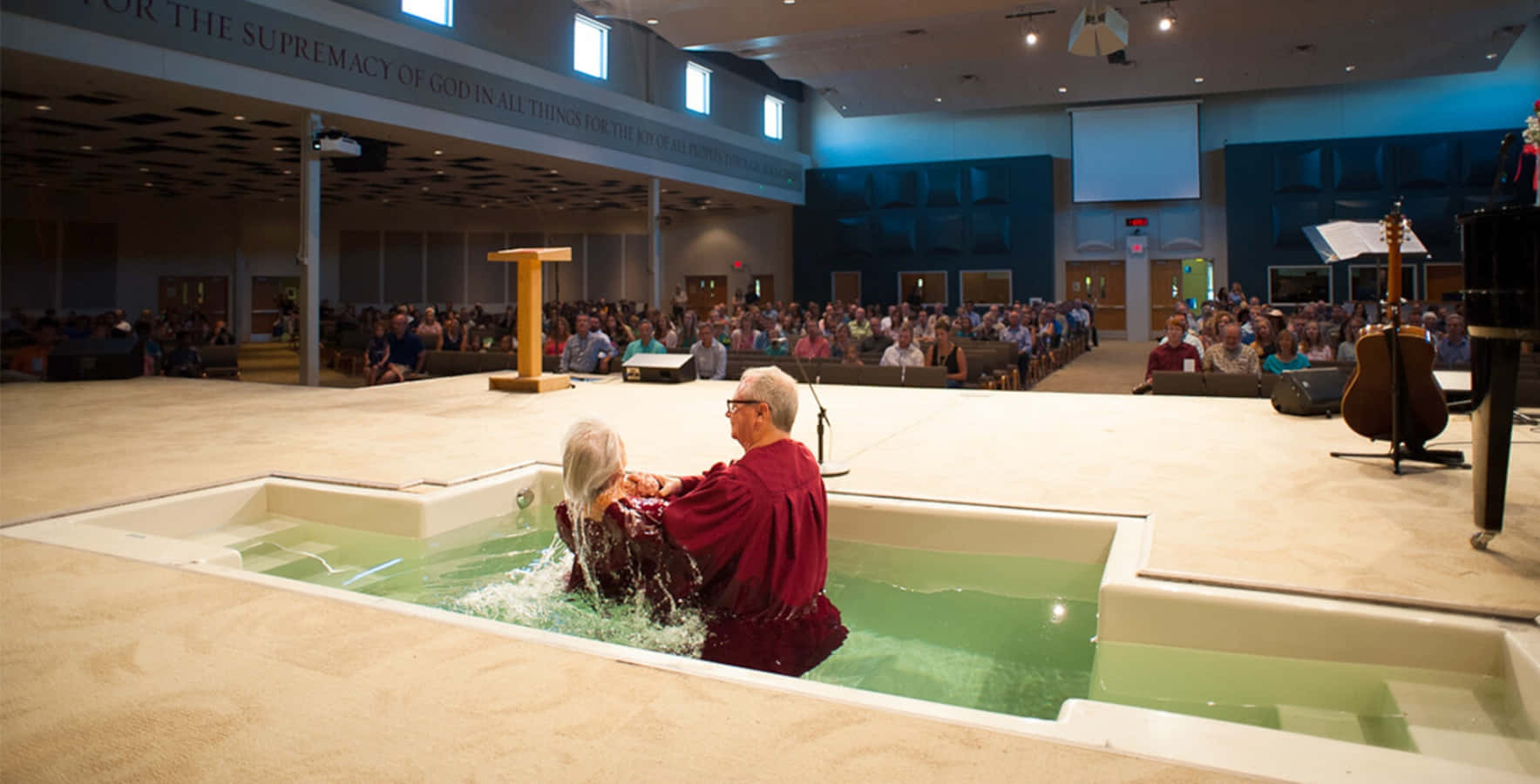 Baptism 桌布 壁紙 — 一位身穿紅色長袍的長者男性在洗禮池中為一位年長女性施洗，會眾在現代教堂的長椅上觀看。