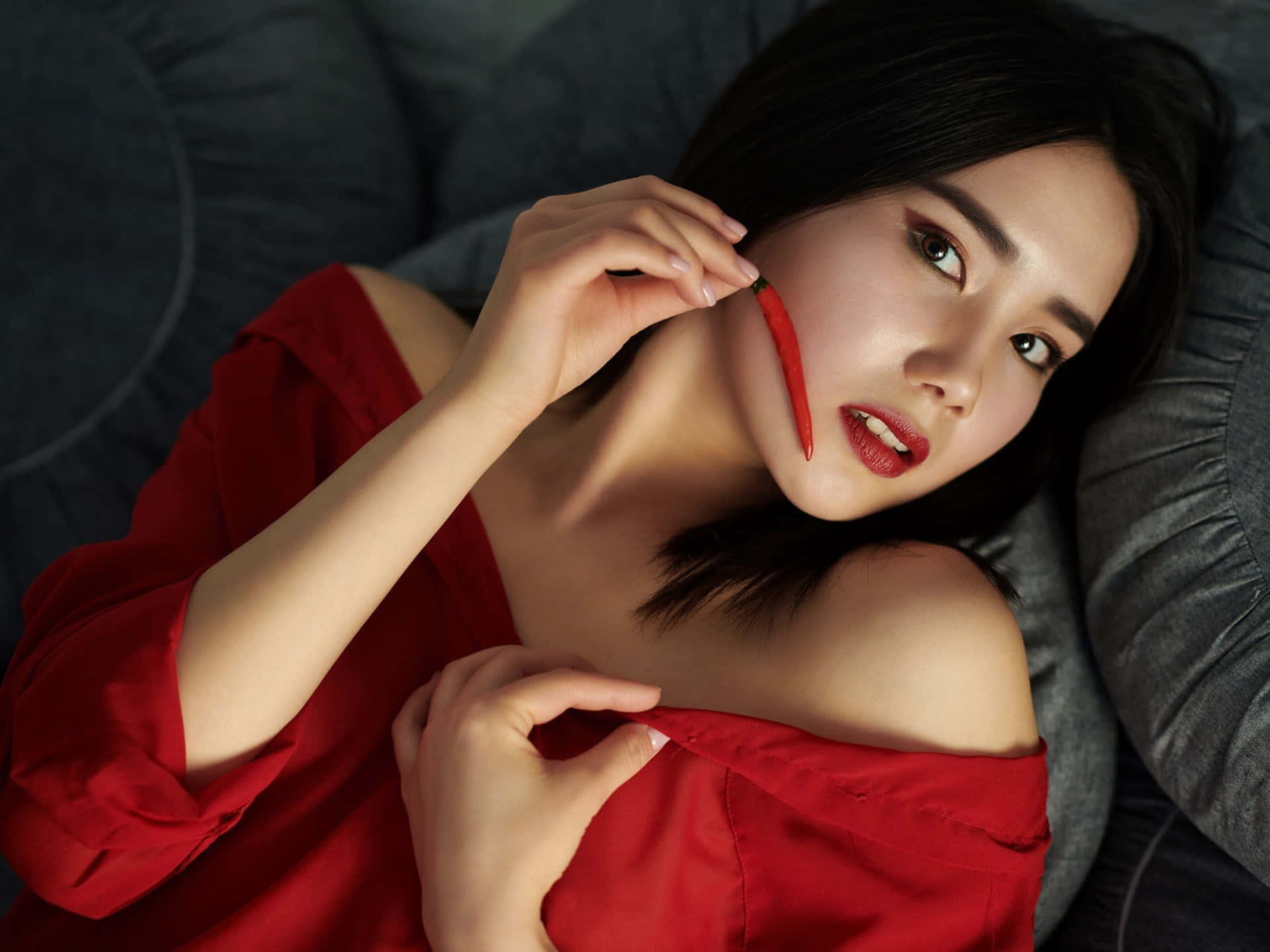 美女 桌布 壁紙 — 一位身穿紅色露肩上衣的女性躺在灰色沙發上，手持紅辣椒靠近臉頰，直視鏡頭。