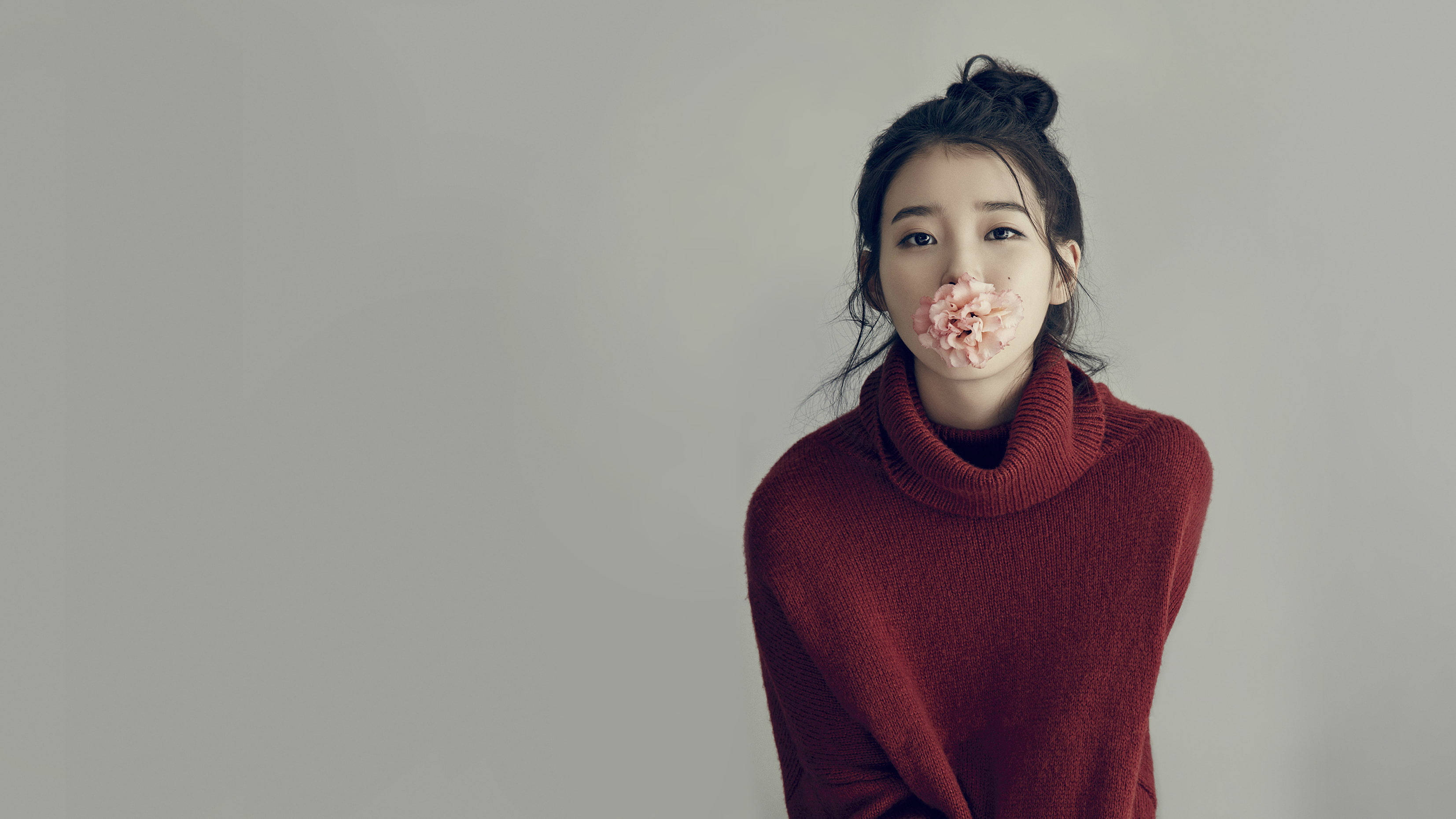 女演員 桌布 壁紙 — 一位身穿紅色高領毛衣的年輕女性，在純灰色背景前，將一朵粉紅色的花放在嘴上。