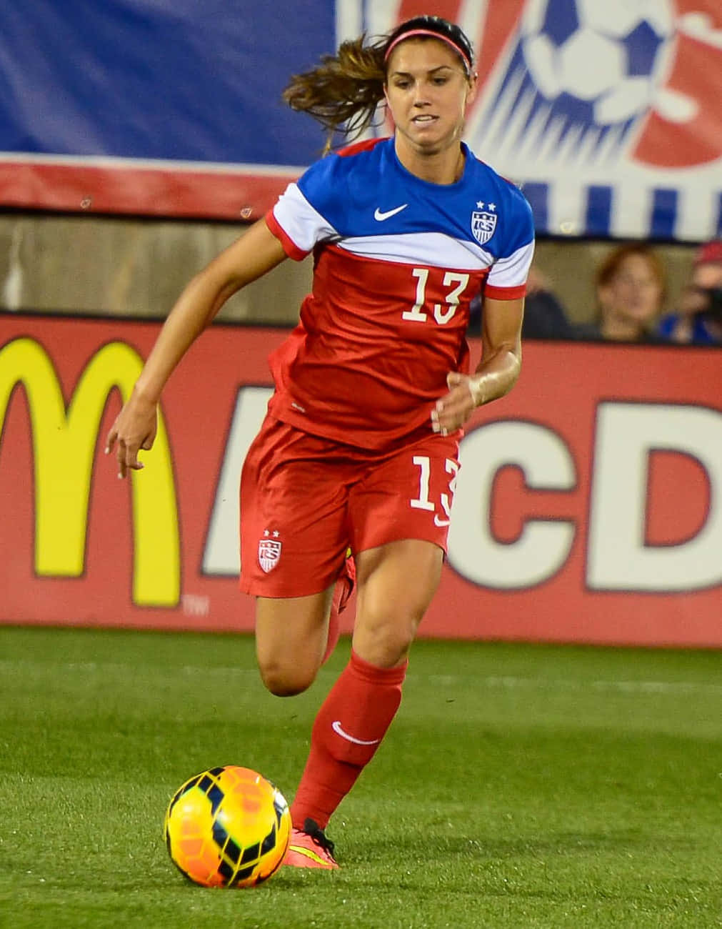 Alex Morgan 桌布 壁紙 — 一位身穿紅藍USA球衣的女足球員，在球場燈光下於綠色場地上盤帶一個鮮豔的橙黑色足球。
