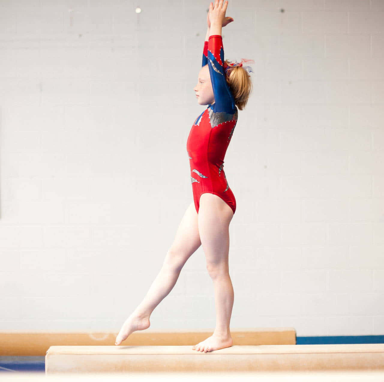 Gymnastics 桌布 壁紙 — 一位身穿紅藍色緊身衣的年輕體操運動員在平衡木上優雅地保持平衡，一條腿伸直，雙臂高舉。