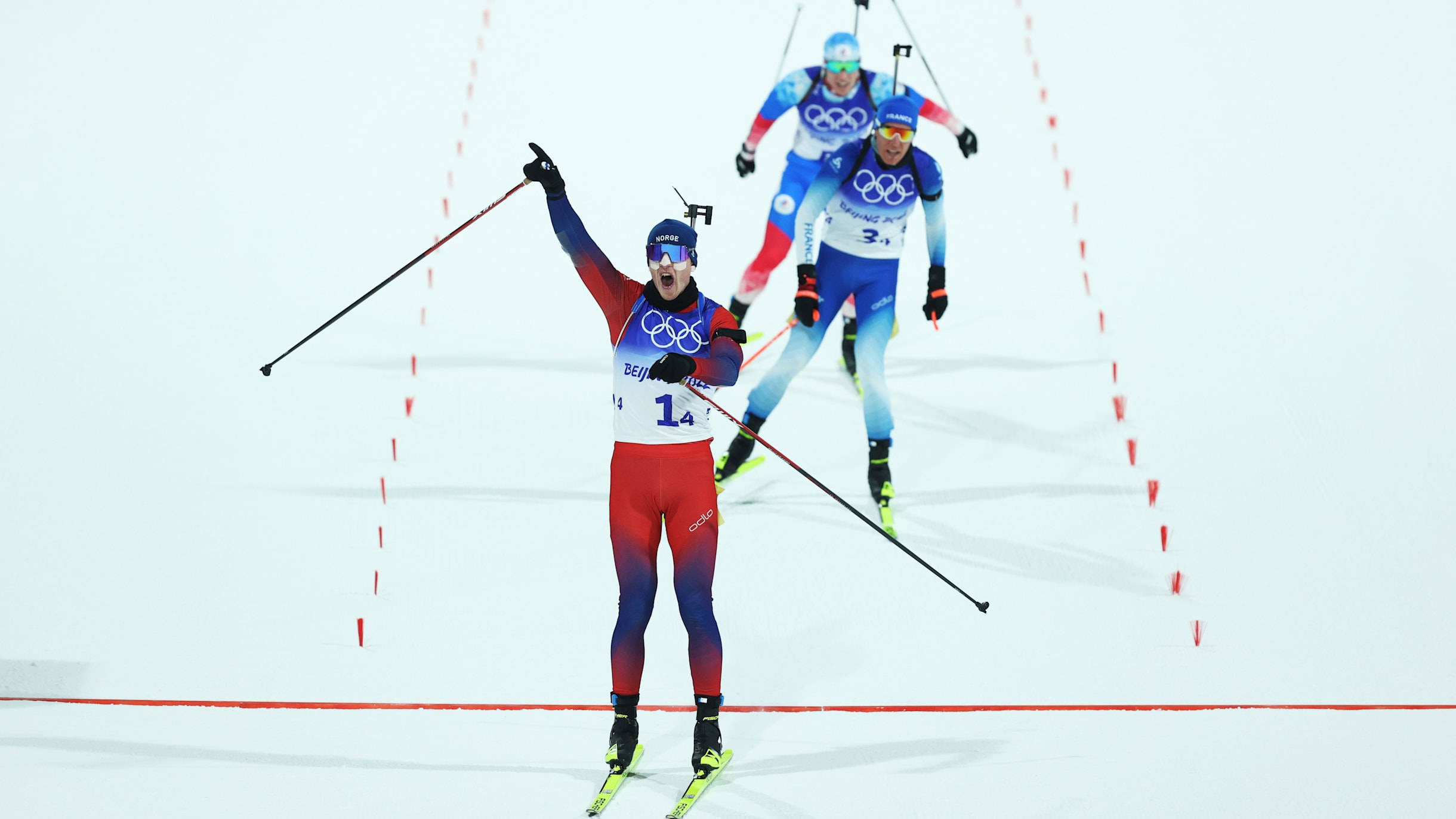 運動 桌布 壁紙 — 一位身穿紅藍裝備的越野滑雪運動員高舉雙臂慶祝衝過終點線，身後有競爭對手在雪地上緊隨。