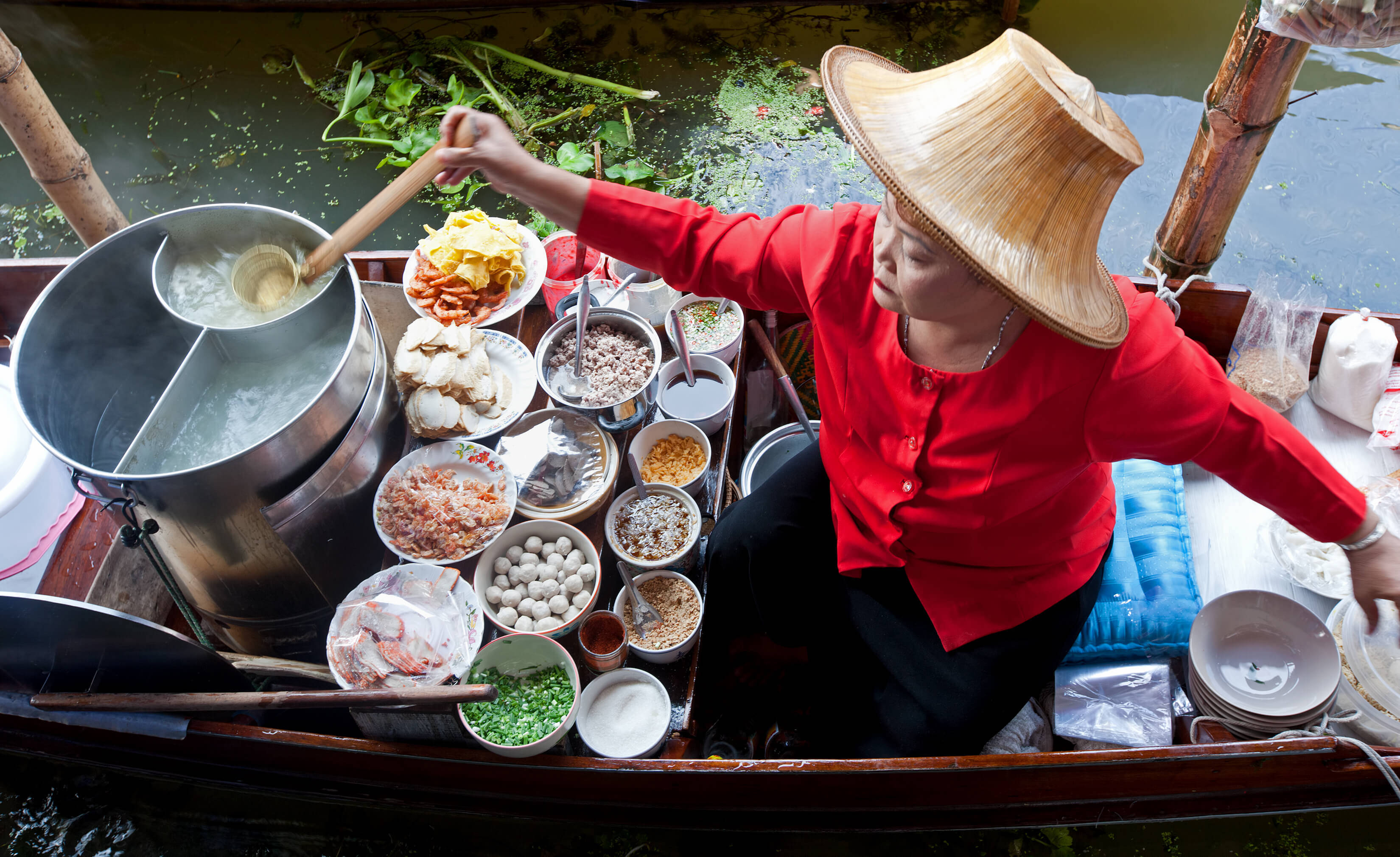 Bangkok 桌布 壁紙 — 一位身穿紅衫、戴草帽的小販在船上攪拌湯底，船上裝滿了盛載街頭小吃食材的碗，位於河上。