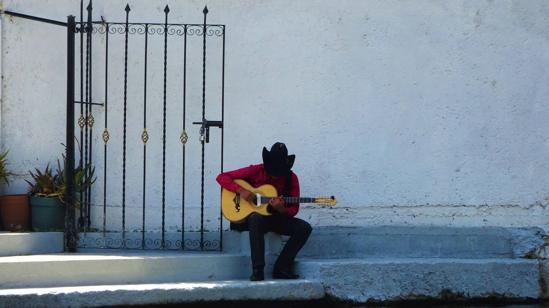 Mexican 桌布 壁紙 — 一位身穿紅衫黑帽的音樂家坐在石階上，旁邊是白色牆壁和華麗大門，彈奏著黃色吉他。
