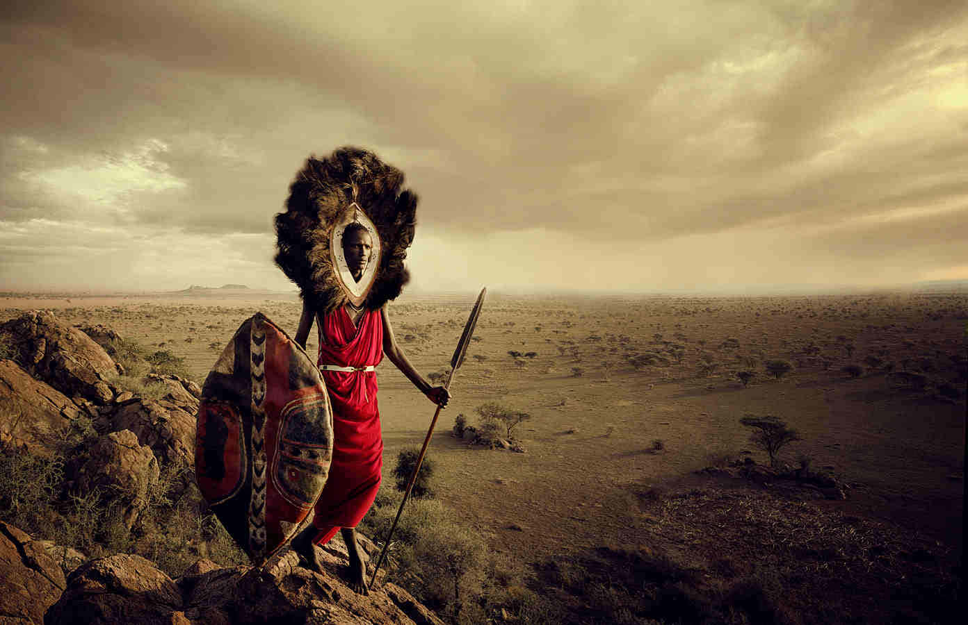 African Culture 桌布 壁紙 — 一位身穿紅袍、頭戴羽毛頭飾的戰士站在岩石露頭上，俯瞰著多雲天空下廣闊乾旱的稀樹草原。