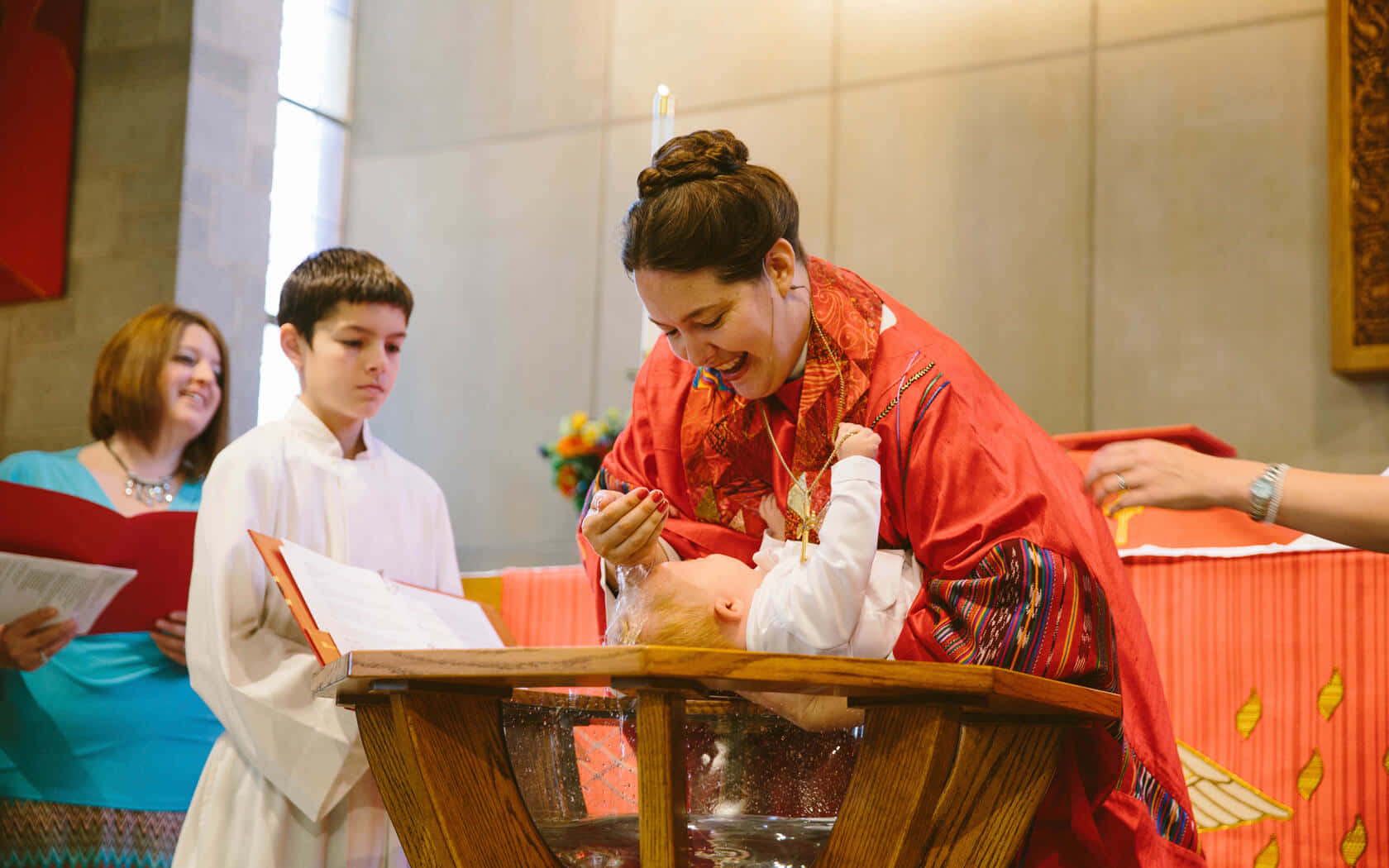 Baptism 桌布 壁紙 — 一位身穿紅袍的喜樂牧師在木製洗禮盆中為嬰兒施洗，一個男孩和一個女人在教堂裡觀看。