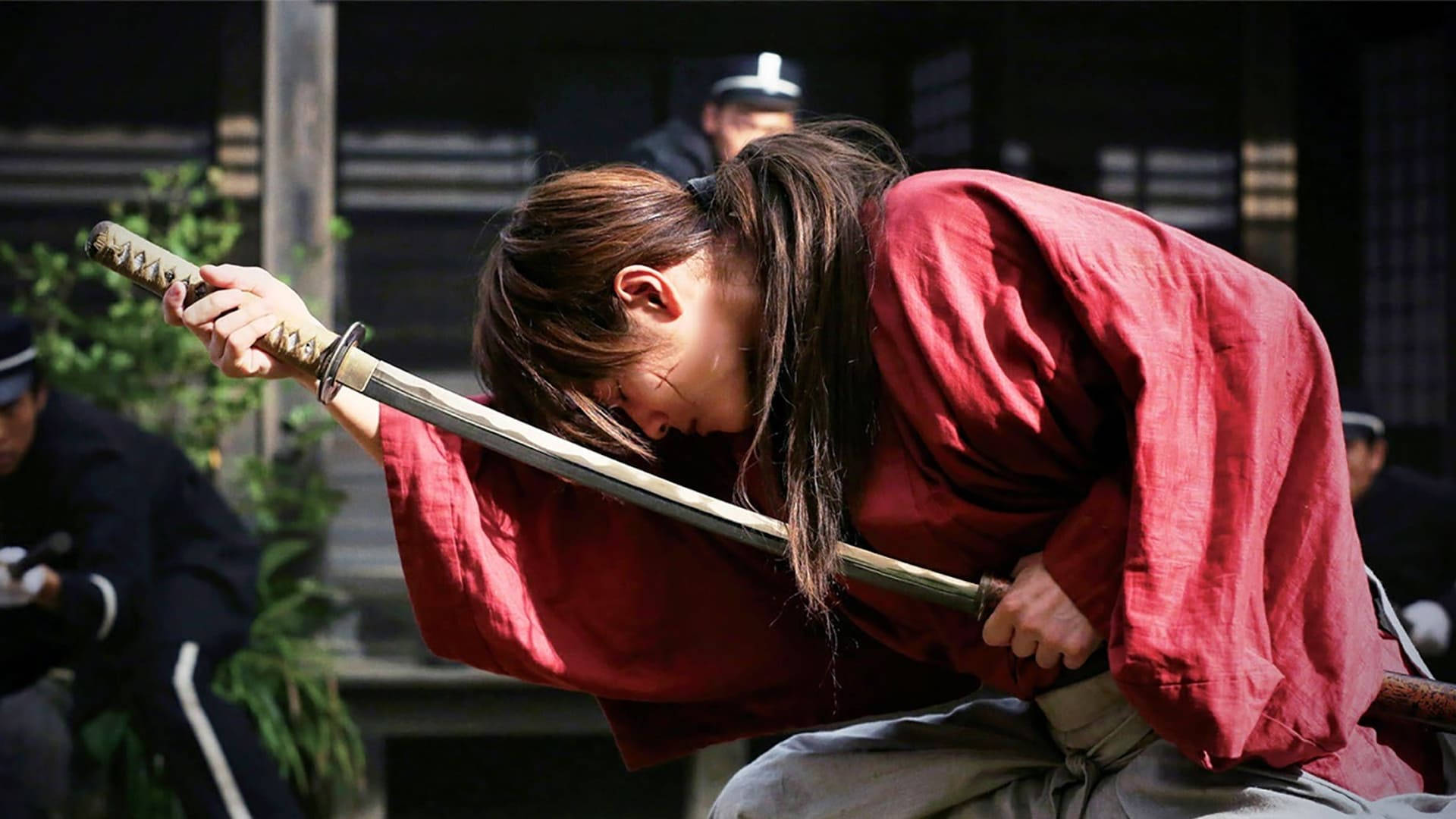 Kenshin 桌布 壁紙 — 一位身穿紅袍的武士在傳統日本庭院中深深鞠躬，同時緊握著一把武士刀。