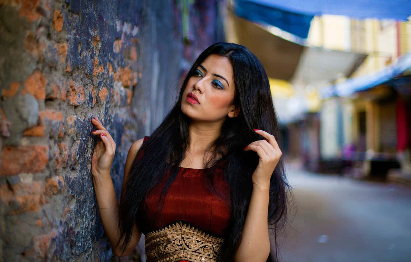 photo 桌布 壁紙 — 一位身穿紅金傳統連衣裙的女性，在狹窄的城市小巷中靠著風化的磚牆。