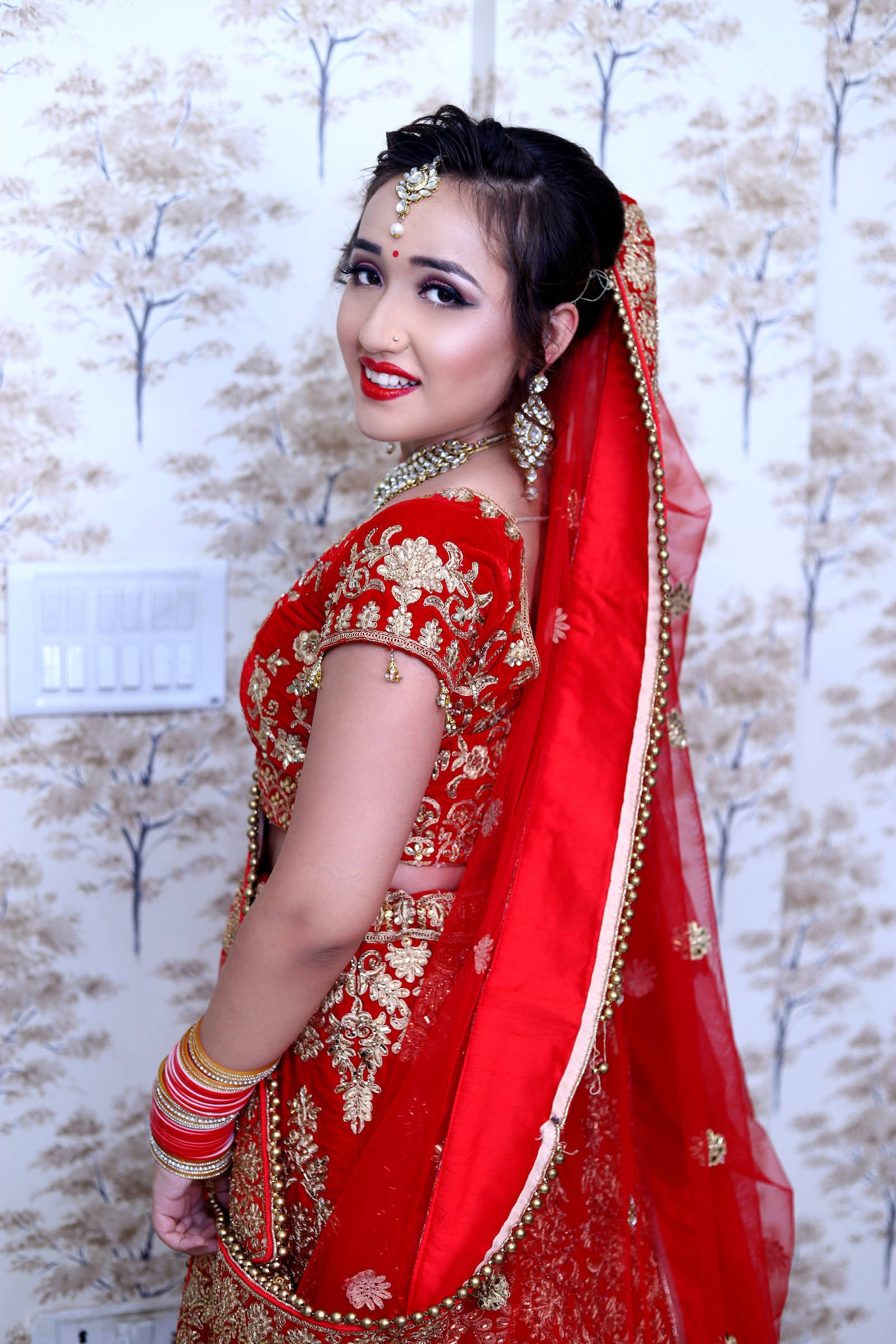 Indian 桌布 壁紙 — 一位身穿紅金刺繡新娘禮服（lehenga）的耀眼女性，在樹紋壁紙前優雅擺姿。
