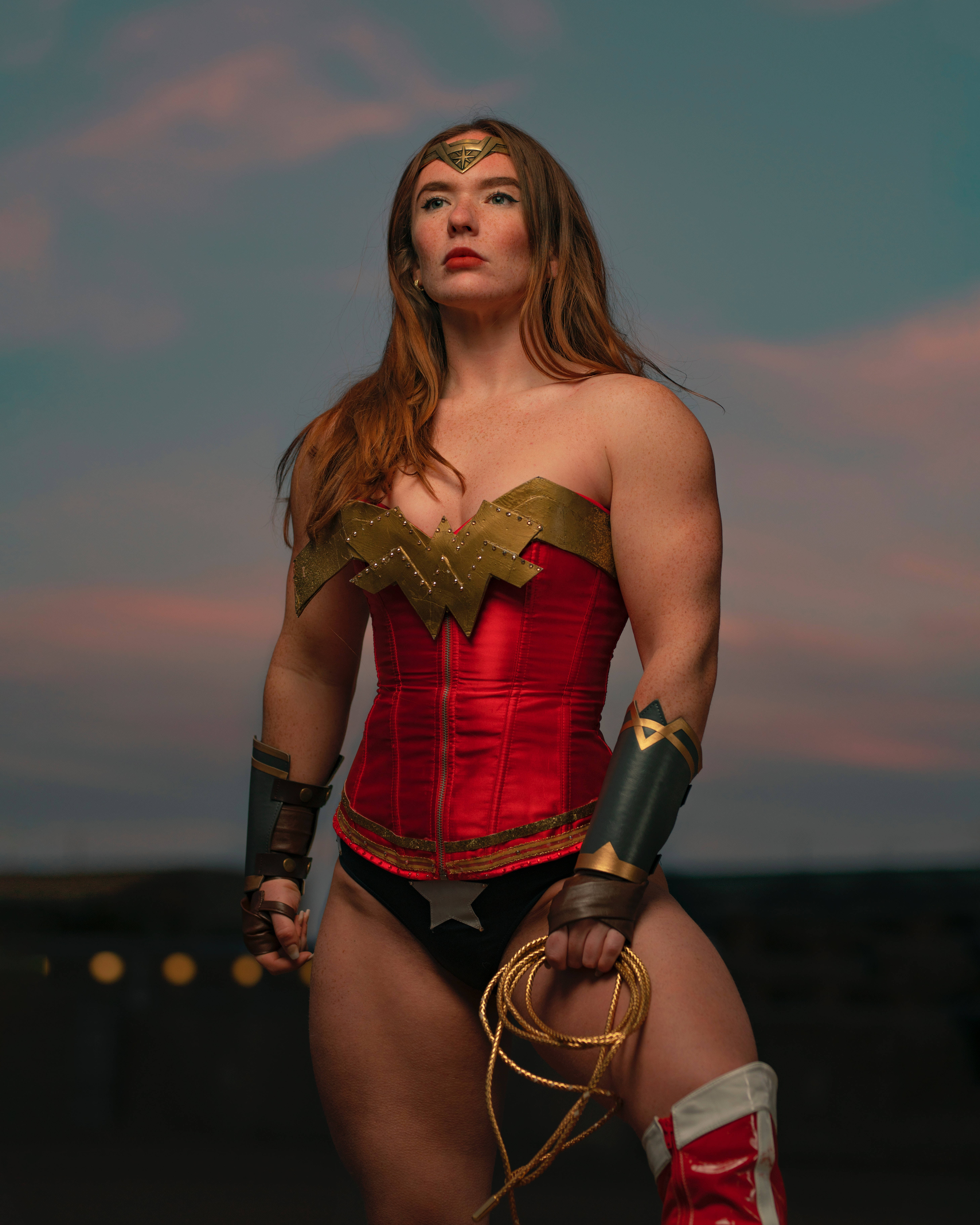 superhero supreme 桌布 壁紙 — 一位身穿紅金相間神力女超人服裝的女性，在暮色天空下自信地站立，手持套索。