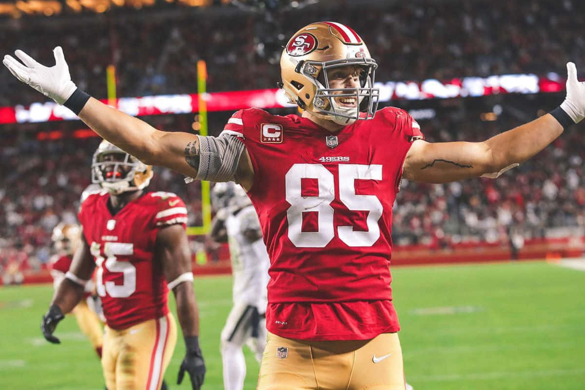 George Kittle 桌布 壁紙 — 一位身穿紅金色85號球衣的舊金山49人隊美式足球員，在夜間燈火通明的球場上張開雙臂慶祝。