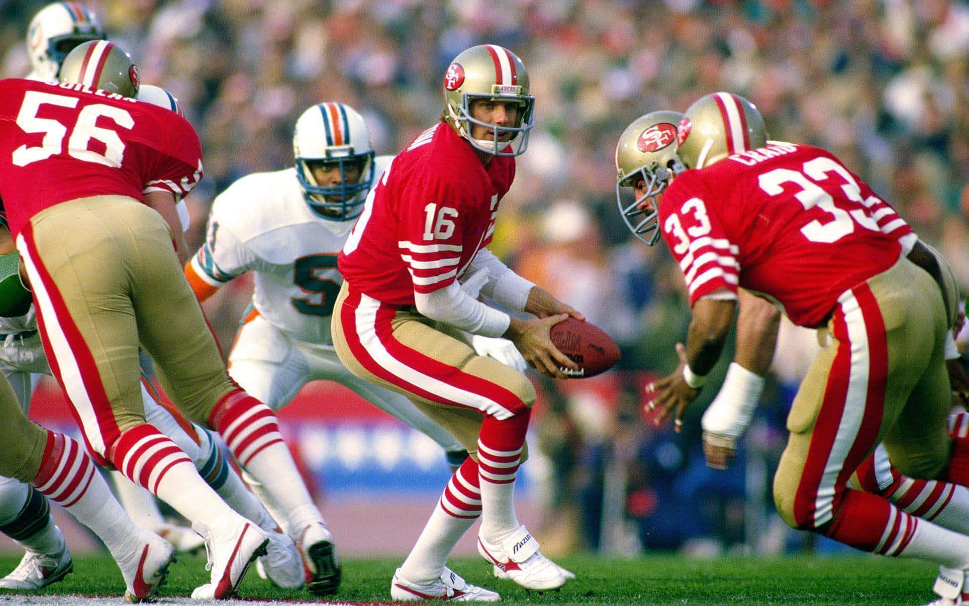 Joe Montana 桌布 壁紙 — 一位身穿紅金色球衣的舊金山四九人四分衛在日間比賽中準備傳球，隊友和對手正在行動。