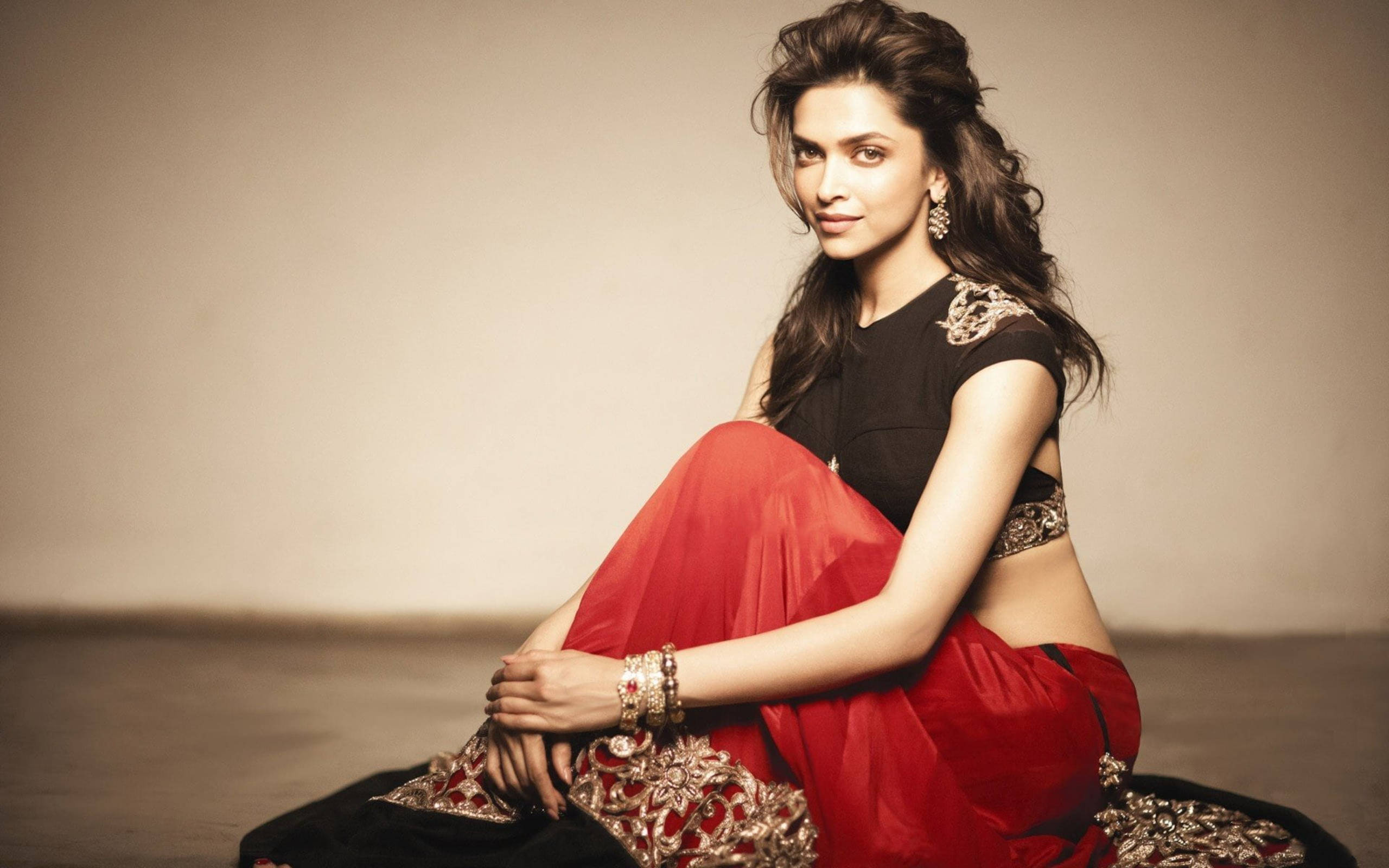 Deepika 桌布 壁紙 — 一位身穿紅黑傳統服飾的女性，佩戴華麗珠寶，在柔和的工作室背景前優雅地坐著。