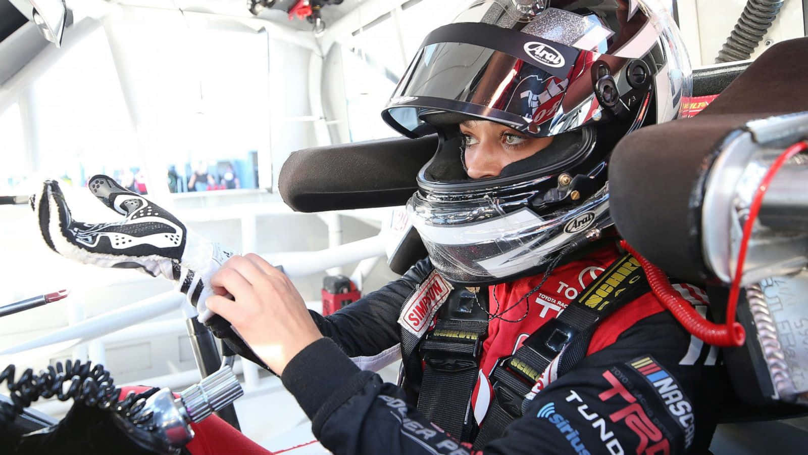 nascar 桌布 壁紙 — 一位身穿紅黑賽車服和頭盔的賽車手正在賽車駕駛艙內調整手套。