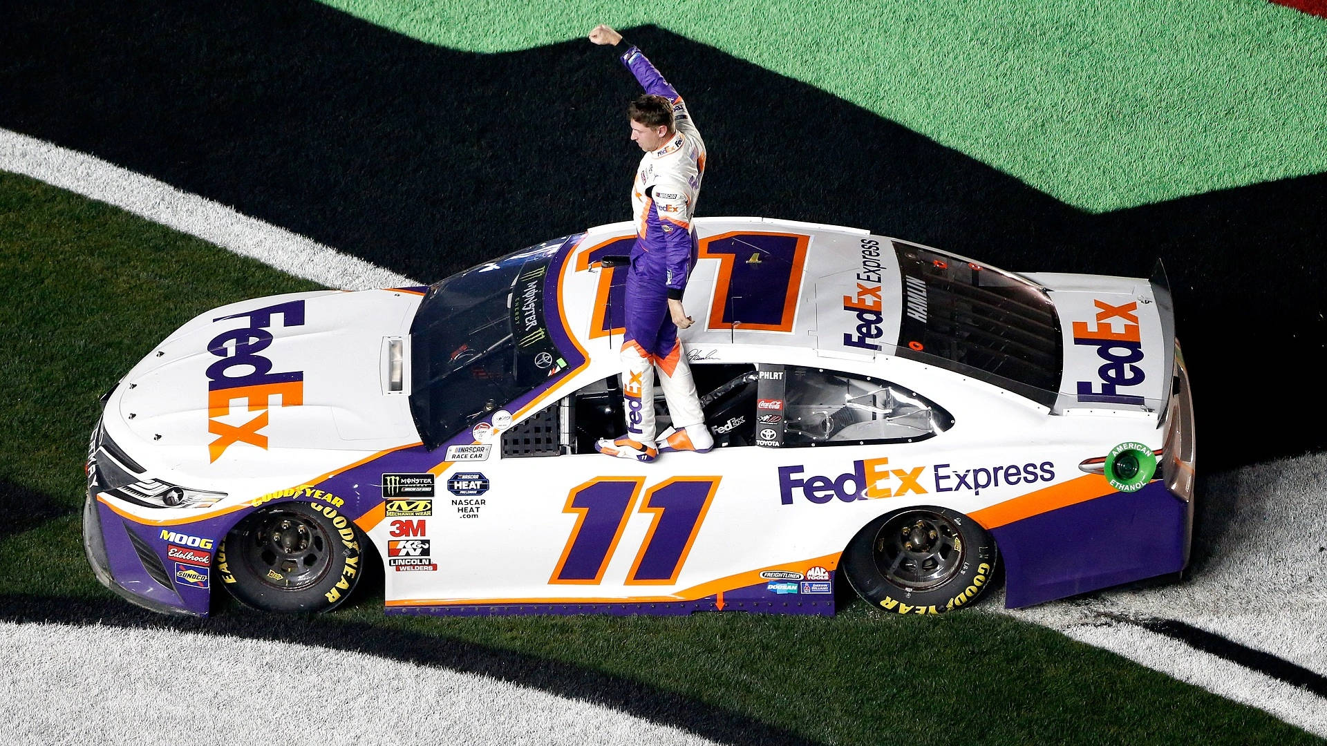 Denny Hamlin 桌布 壁紙 — 一位身穿紫白色的NASCAR車手在夜間的方格賽道上，站在他的FedEx Express賽車頂上慶祝勝利。