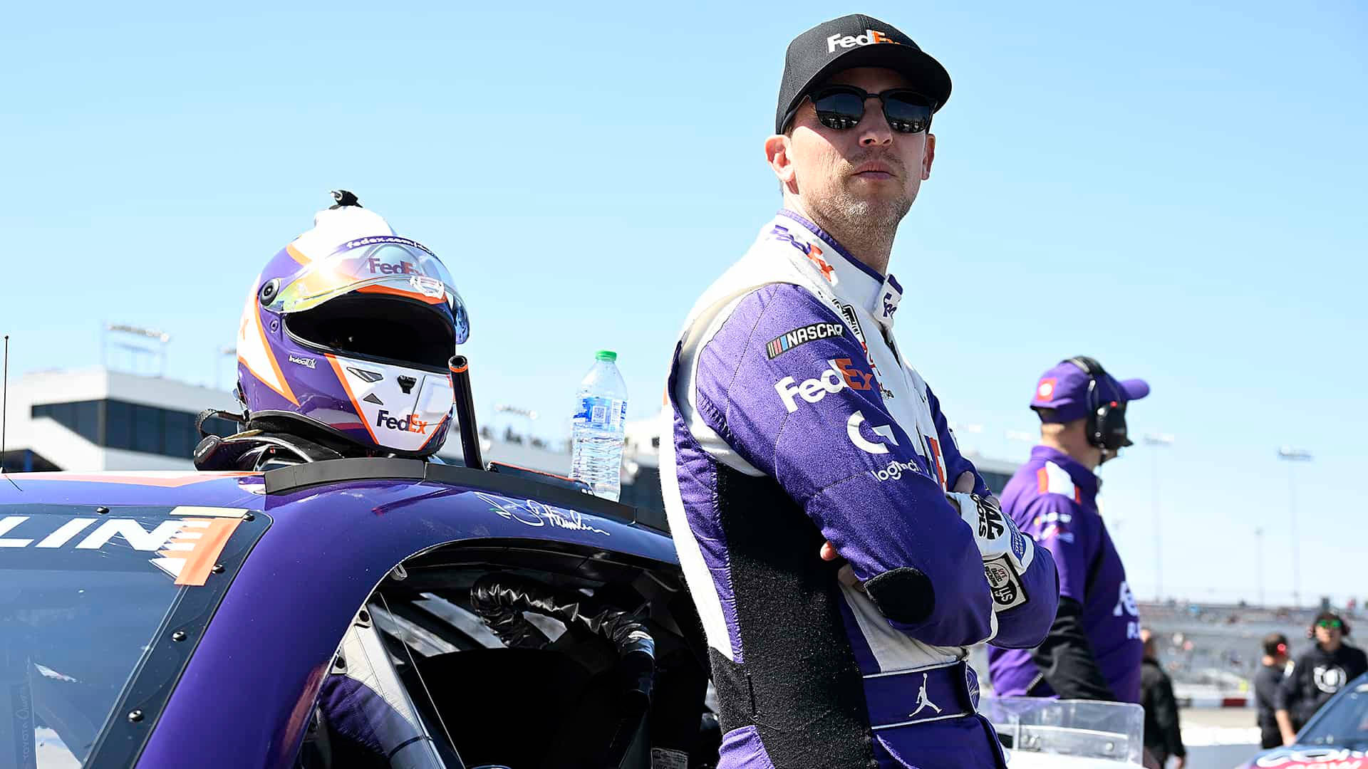 Denny Hamlin 桌布 壁紙 — 一位身穿紫色FedEx制服的NASCAR車手在明亮的日光下自信地站在他的賽車旁邊。