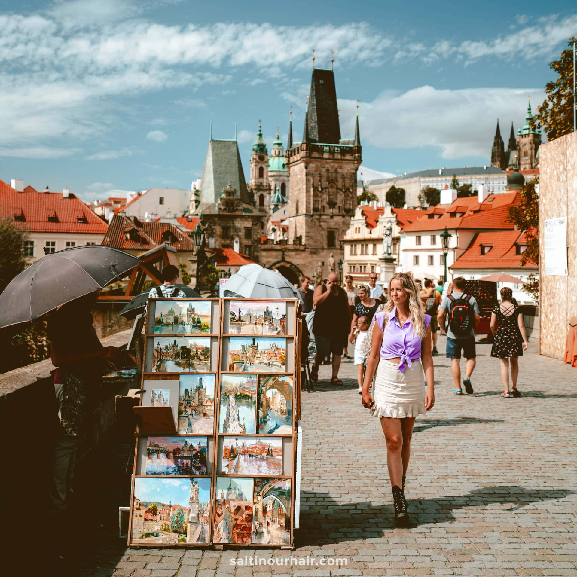 Europe 桌布 壁紙 — 一位身穿紫色上衣的女子在陽光明媚的鵝卵石橋上走過街頭藝術家的展覽，背景是歷史悠久的塔樓和紅色屋頂。