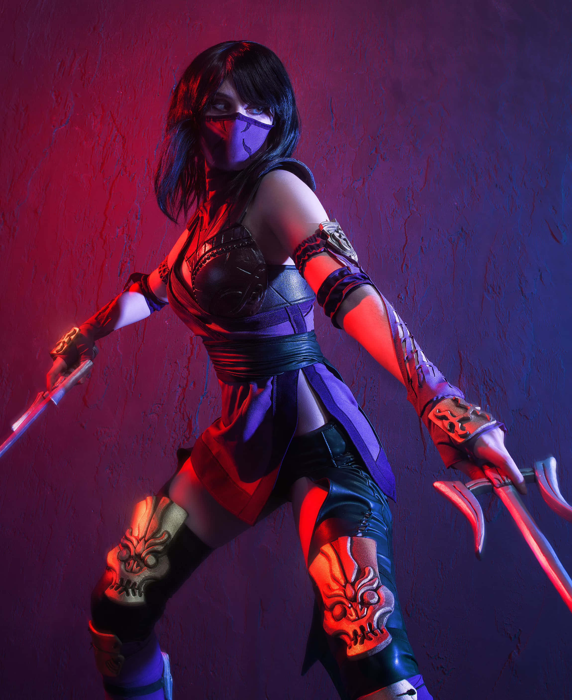 Mortal Kombat Cosplays 桌布 壁紙 — 一位身穿紫色和黑色裝備的忍者戰士，在戲劇性的紅色和藍色燈光下，對著有紋理的牆壁揮舞雙刀。