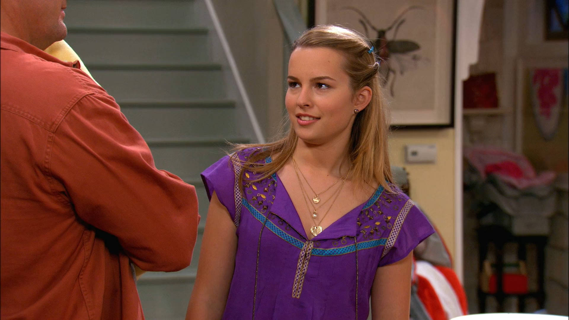 Good Luck Charlie 桌布 壁紙 — 一位身穿紫色圖案上衣的年輕女性微笑著站在溫暖照明的室內環境中，身後有樓梯和裱框藝術品。