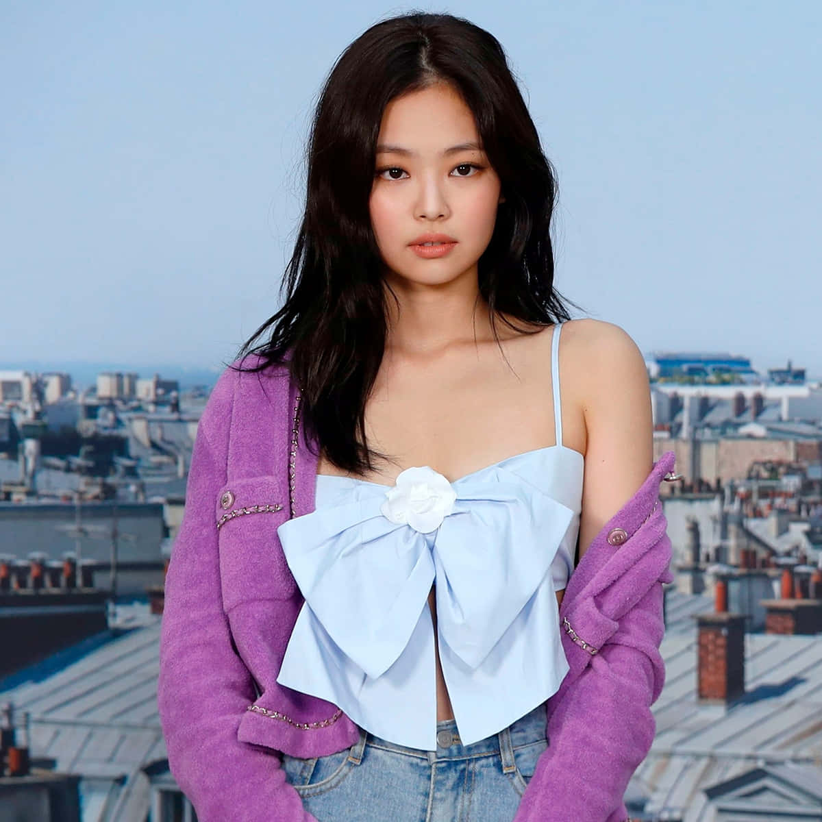 BLACKPINK 桌布 壁紙 — 一位身穿紫色外套和淺藍色蝴蝶結上衣的年輕女性自信地站在城市天際線背景下。