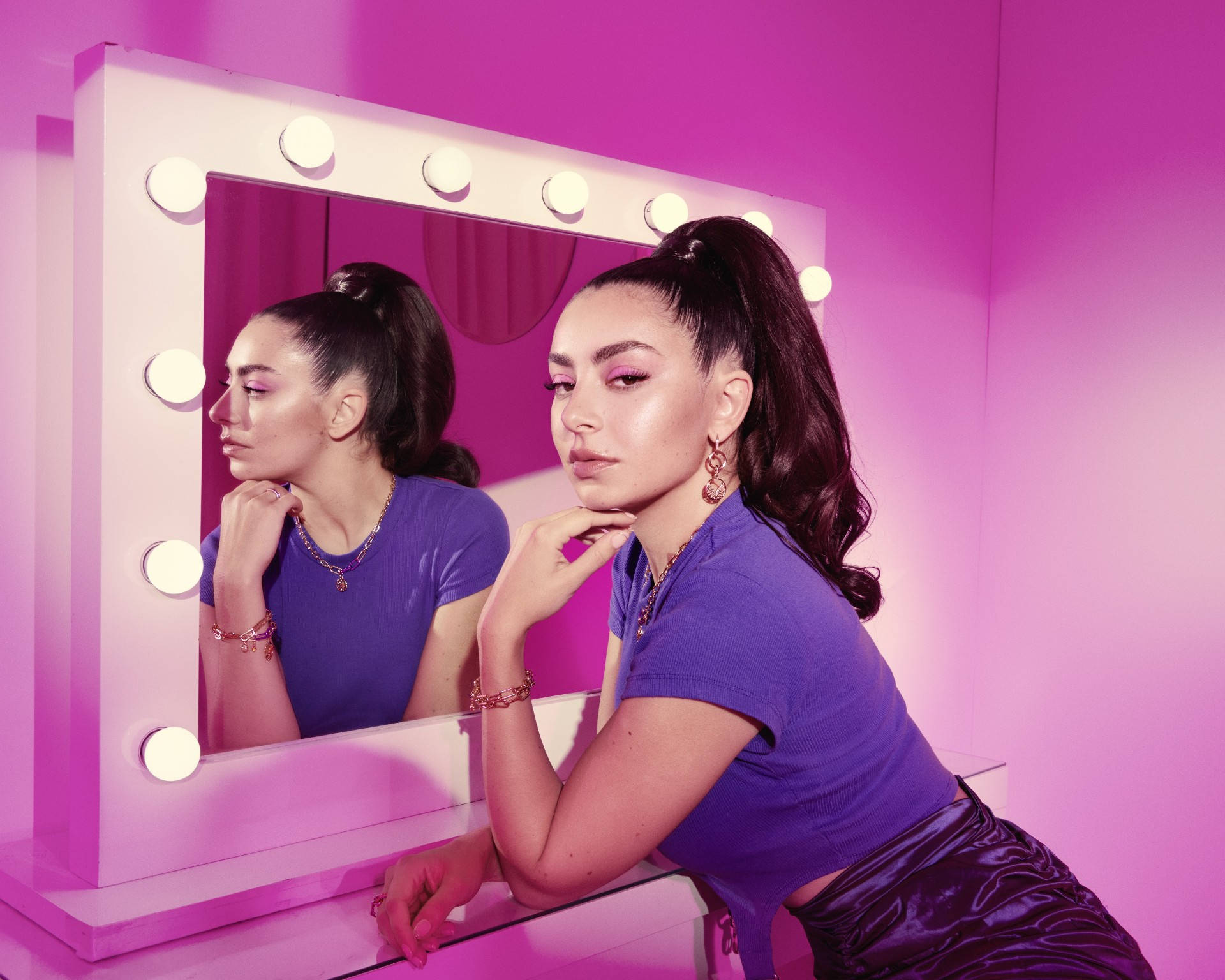 Xcx 桌布 壁紙 — 一位身穿紫色服裝的女性，若有所思地凝視著自己在粉紅色燈光、帶有發光燈泡的梳妝鏡中的倒影。