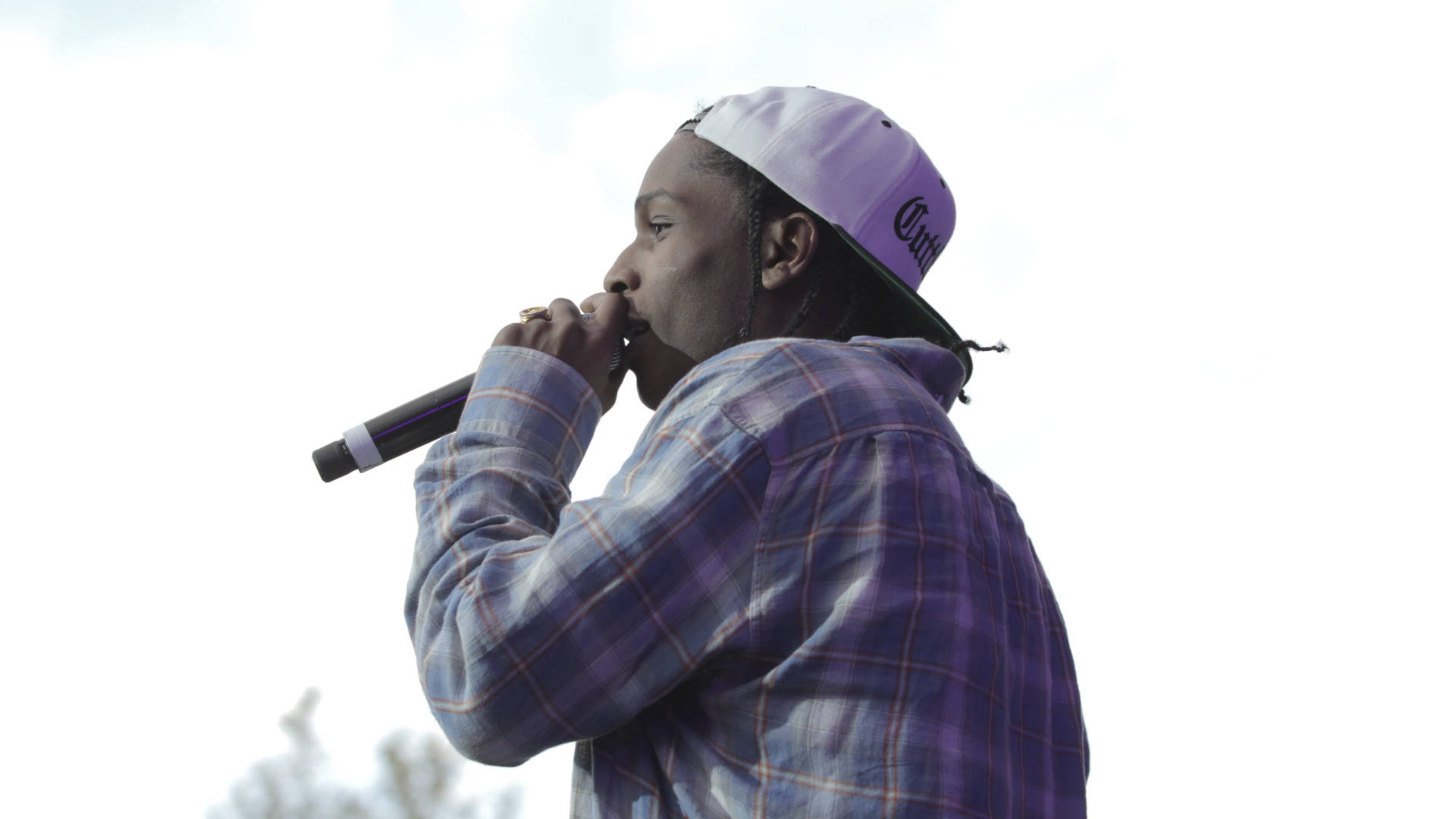 rocky 桌布 壁紙 — 一位身穿紫色格紋襯衫、戴著反戴帽子的饒舌歌手，在明亮的天空下對著麥克風饒舌。