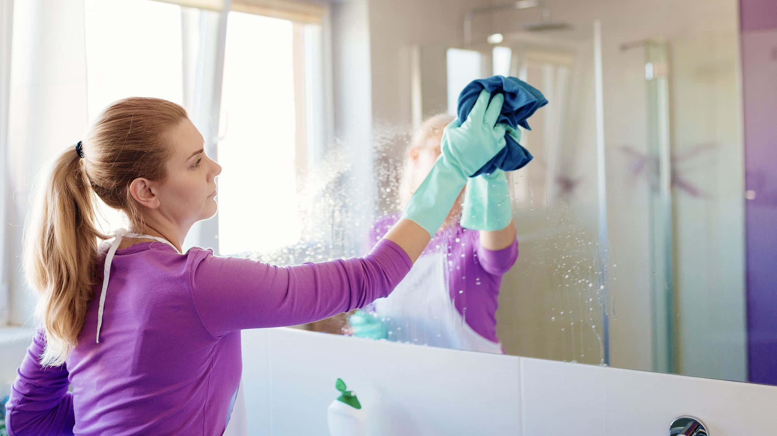 Cleaning 桌布 壁紙 — 一位身穿紫色襯衫和綠色手套的女性，用藍色布噴灑和擦拭浴室鏡子。