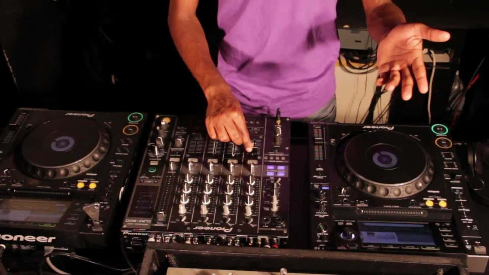 Music 桌布 壁紙 — 一位身穿紫色襯衫的DJ在昏暗的舞台燈光下，於黑色Pioneer唱盤上混音曲目。