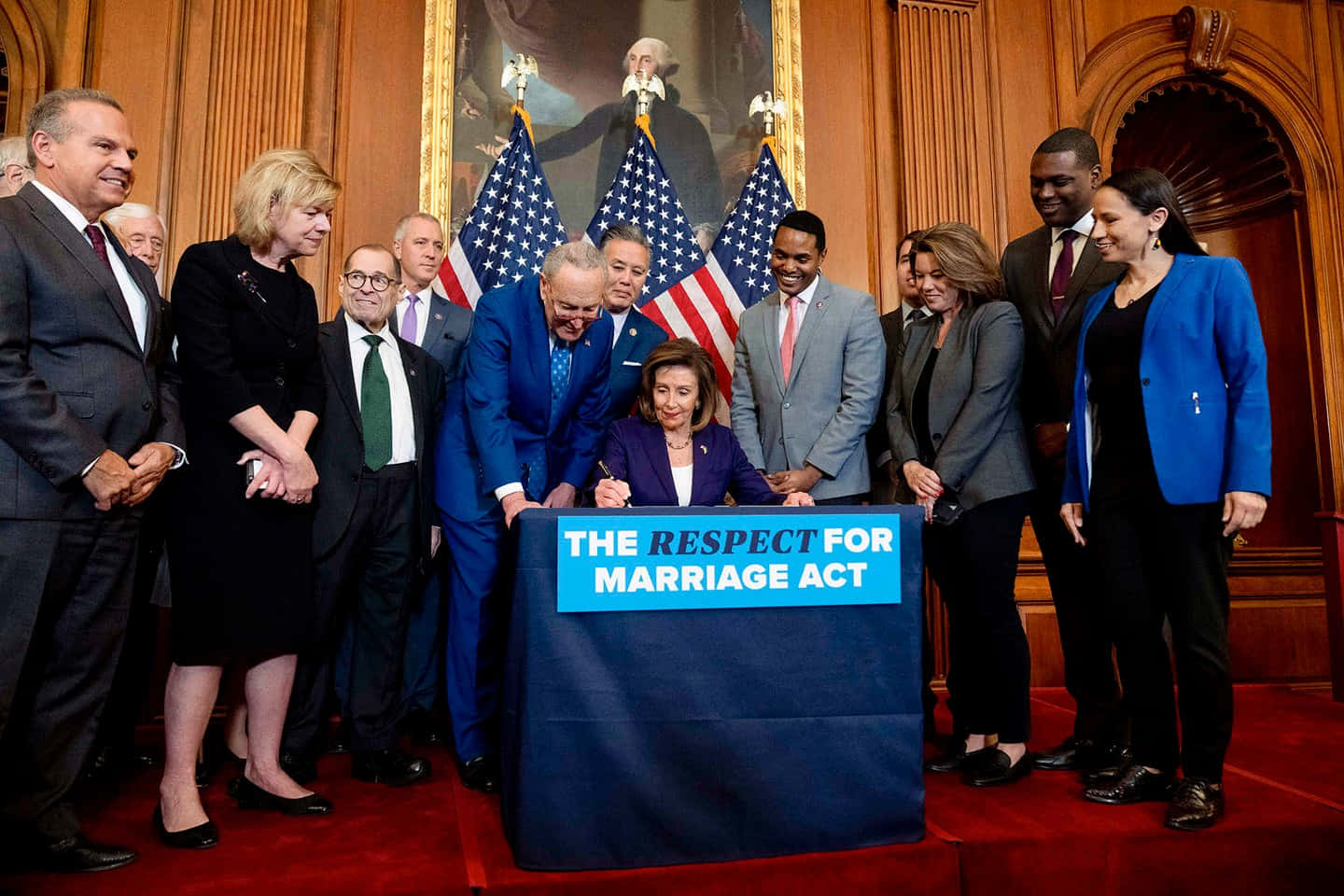 Nancy Pelosi 桌布 壁紙 — 一位身穿紫色西裝的女性在藍色講台上簽署《尊重婚姻法案》，周圍是微笑的立法者，身處正式的木鑲板房間，掛有美國國旗。
