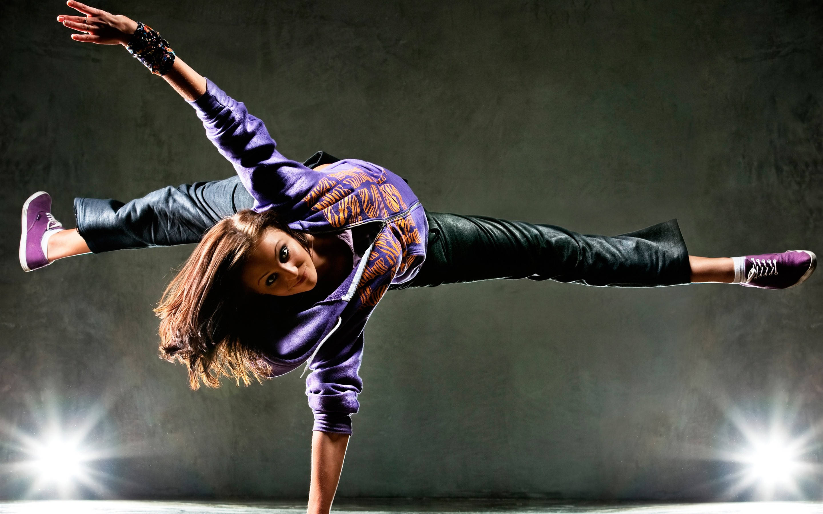 dancer 桌布 壁紙 — 一位身穿紫色連帽衫和黑色褲子的女霹靂舞者在明亮的舞台燈光下，以深灰色牆壁為背景，做出動態的倒立姿勢。