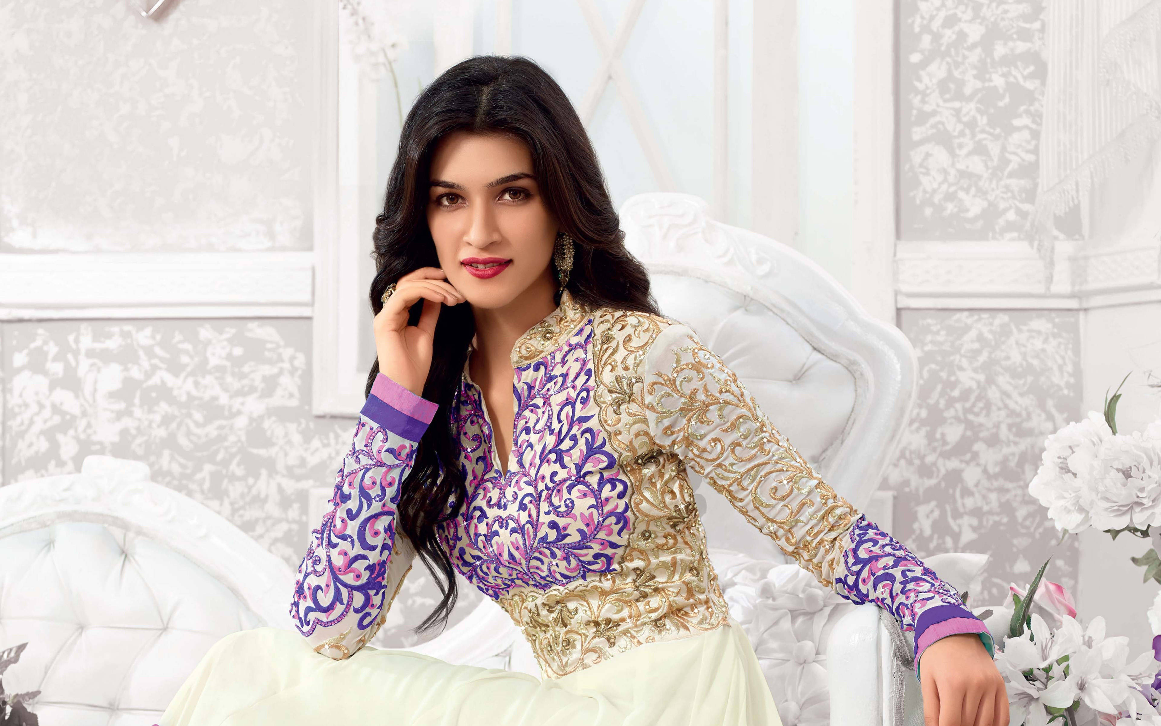 Kriti 桌布 壁紙 — 一位身穿紫金色刺繡服裝的女性優雅地坐在白色簇絨椅上，旁邊有白色花朵。