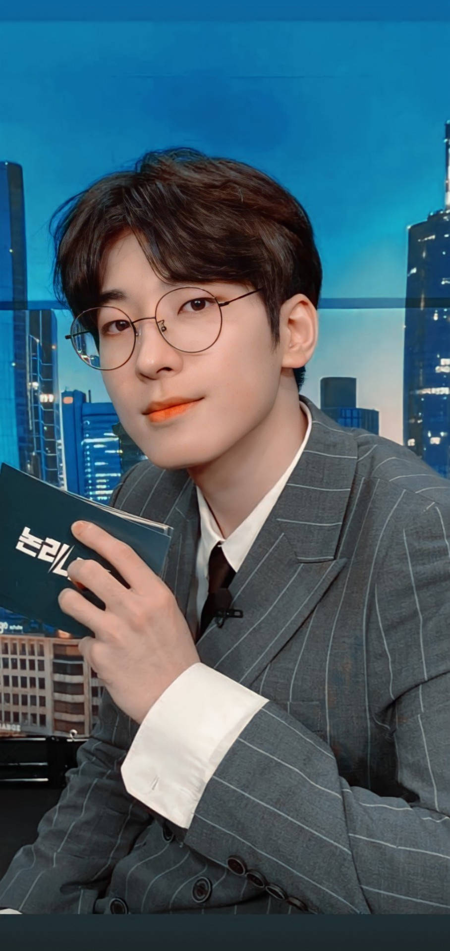 원우 桌布 壁紙 — 一位身穿細條紋西裝、戴眼鏡的年輕男子，以充滿活力的藍色城市景觀為背景，手持一本書。