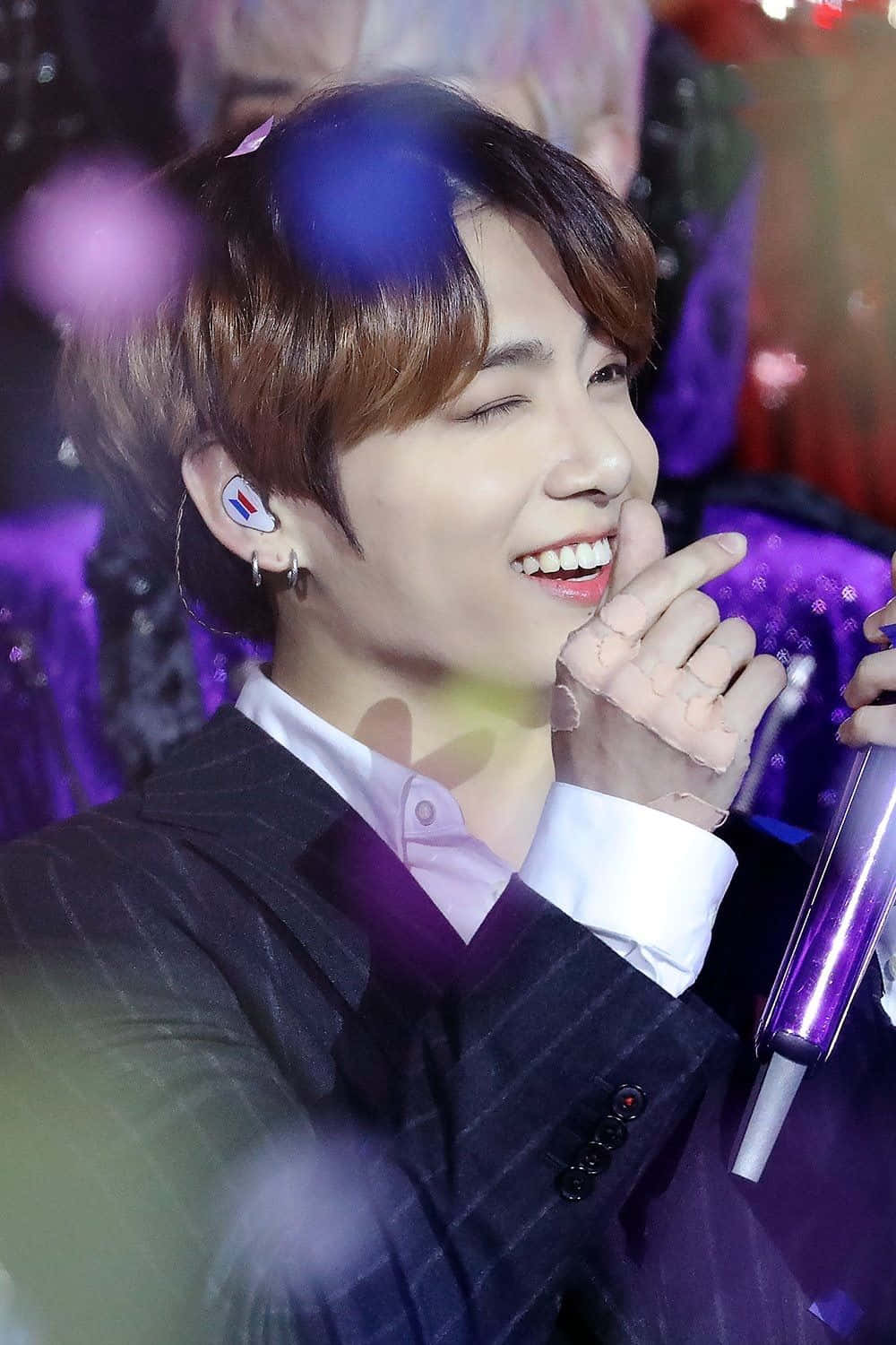 BTS 桌布 壁紙 — 一位身穿細條紋西裝的表演者微笑著眨眼，在舞台燈光下拿著發光的紫色麥克風。