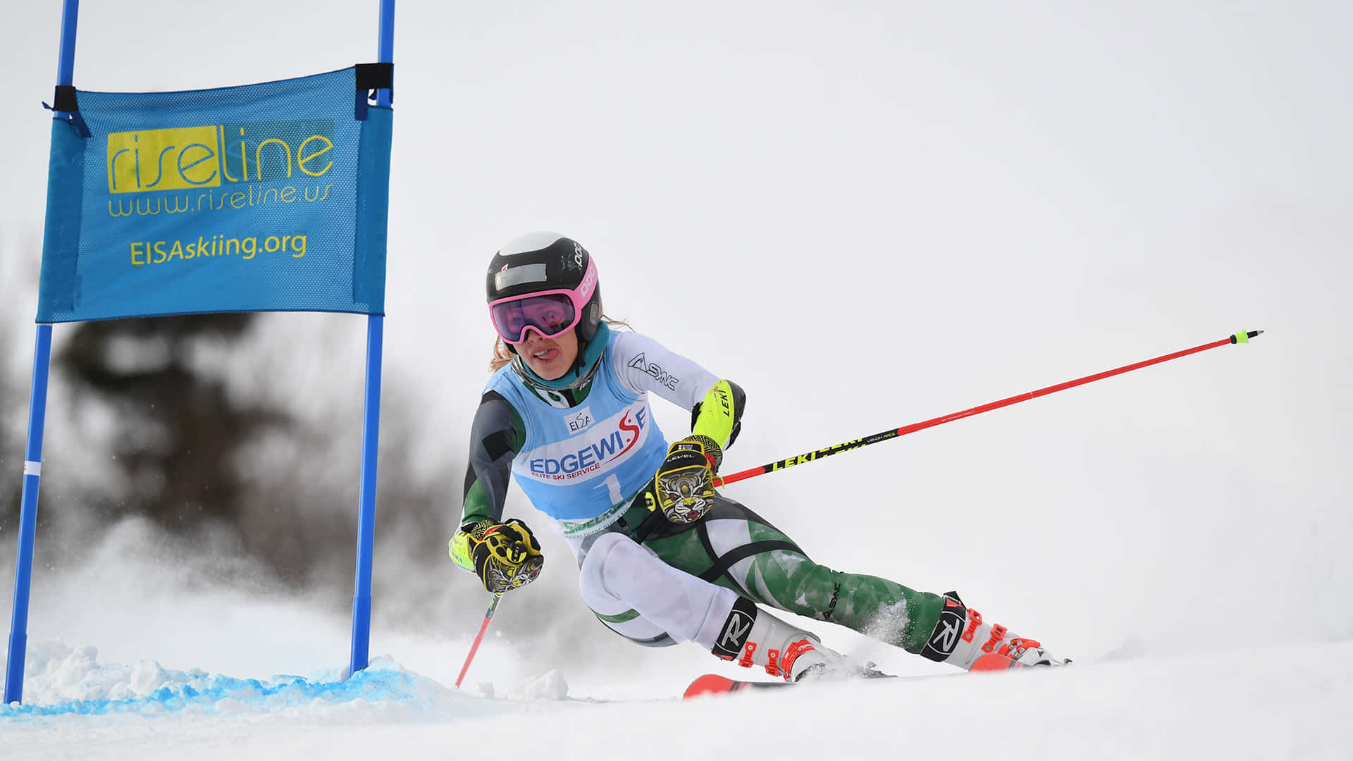 ski 桌布 壁紙 — 一位身穿綠色和白色裝備的女滑雪運動員在比賽中繞過藍色旗門，在雪地上劃出弧線，揚起粉雪。