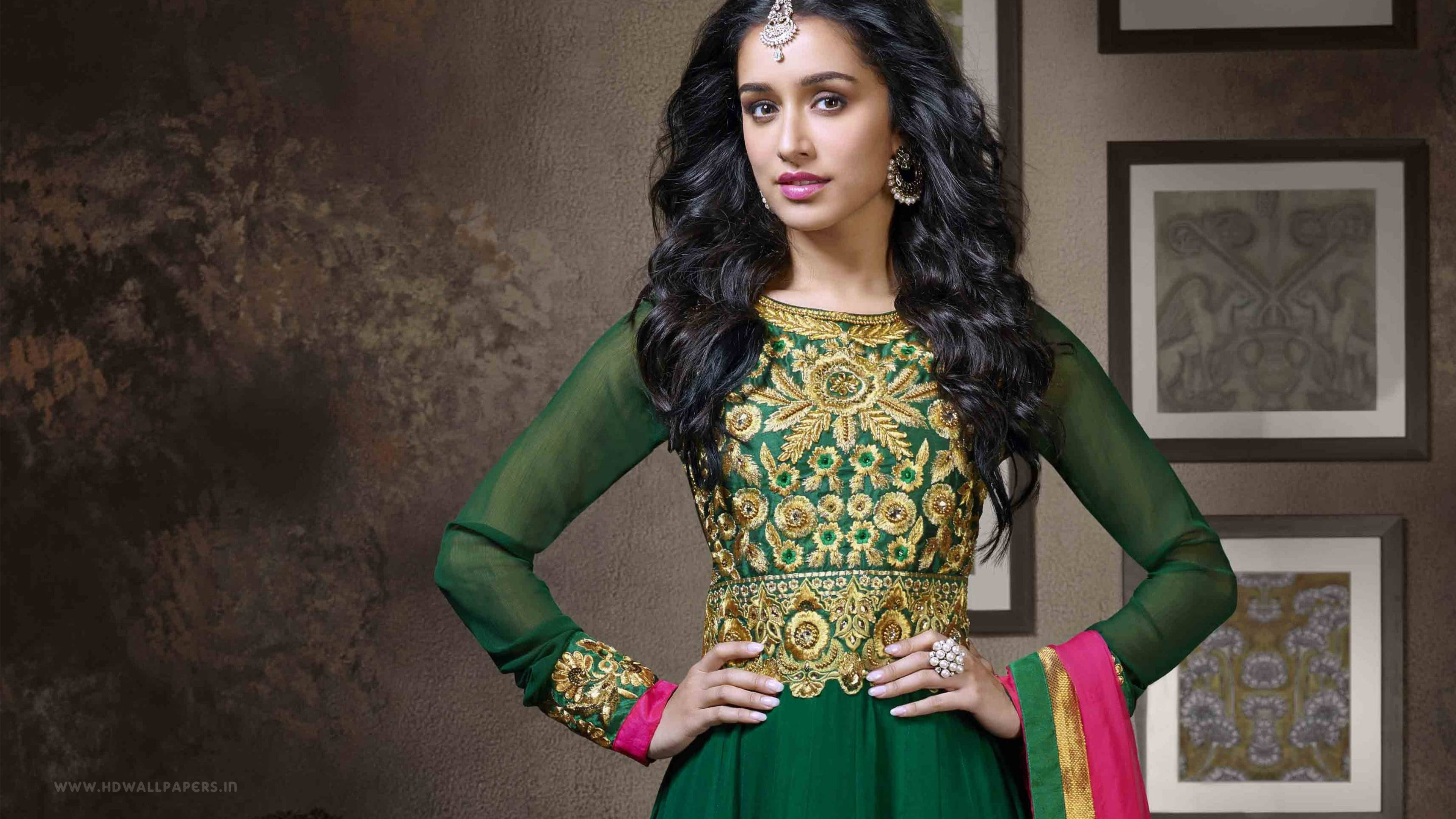 Shraddha Kapoor 桌布 壁紙 — 一位身穿綠色和金色刺繡傳統服裝的女性，自信地在帶有紋理牆壁和裱框藝術品前擺姿勢。