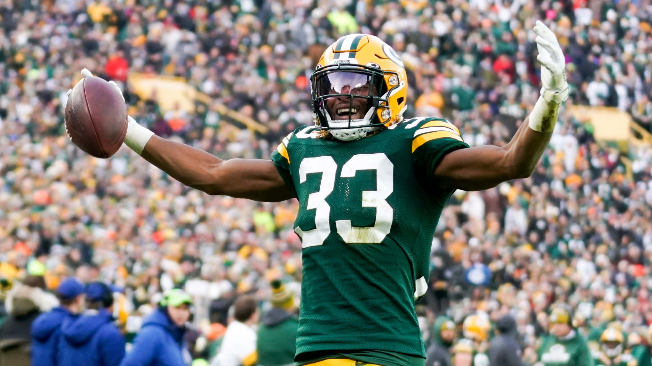 Aaron Jones 桌布 壁紙 — 一位身穿綠色和金色球衣的綠灣包裝工美式足球員，高舉雙臂拿著足球，勝利地慶祝。