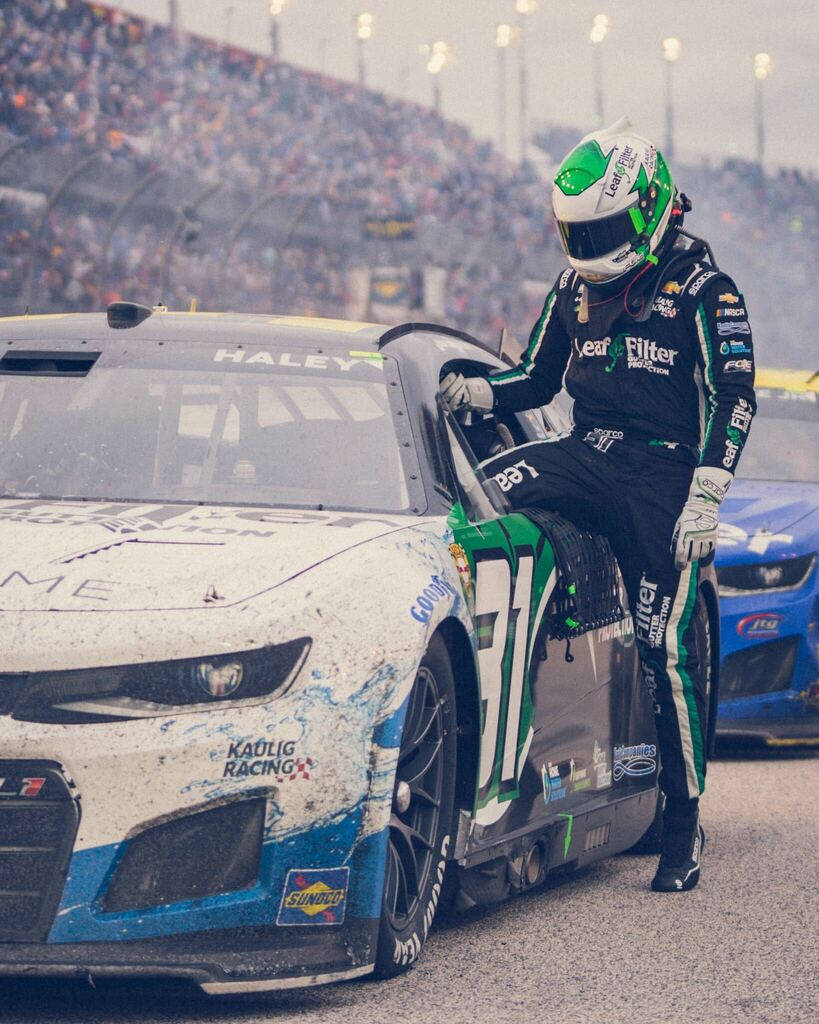 人物 桌布 壁紙 — 一位身穿綠色和黑色裝備的NASCAR車手在擁擠的賽道上靠在他那濺滿泥巴的#11 Kaufig Racing賽車上。